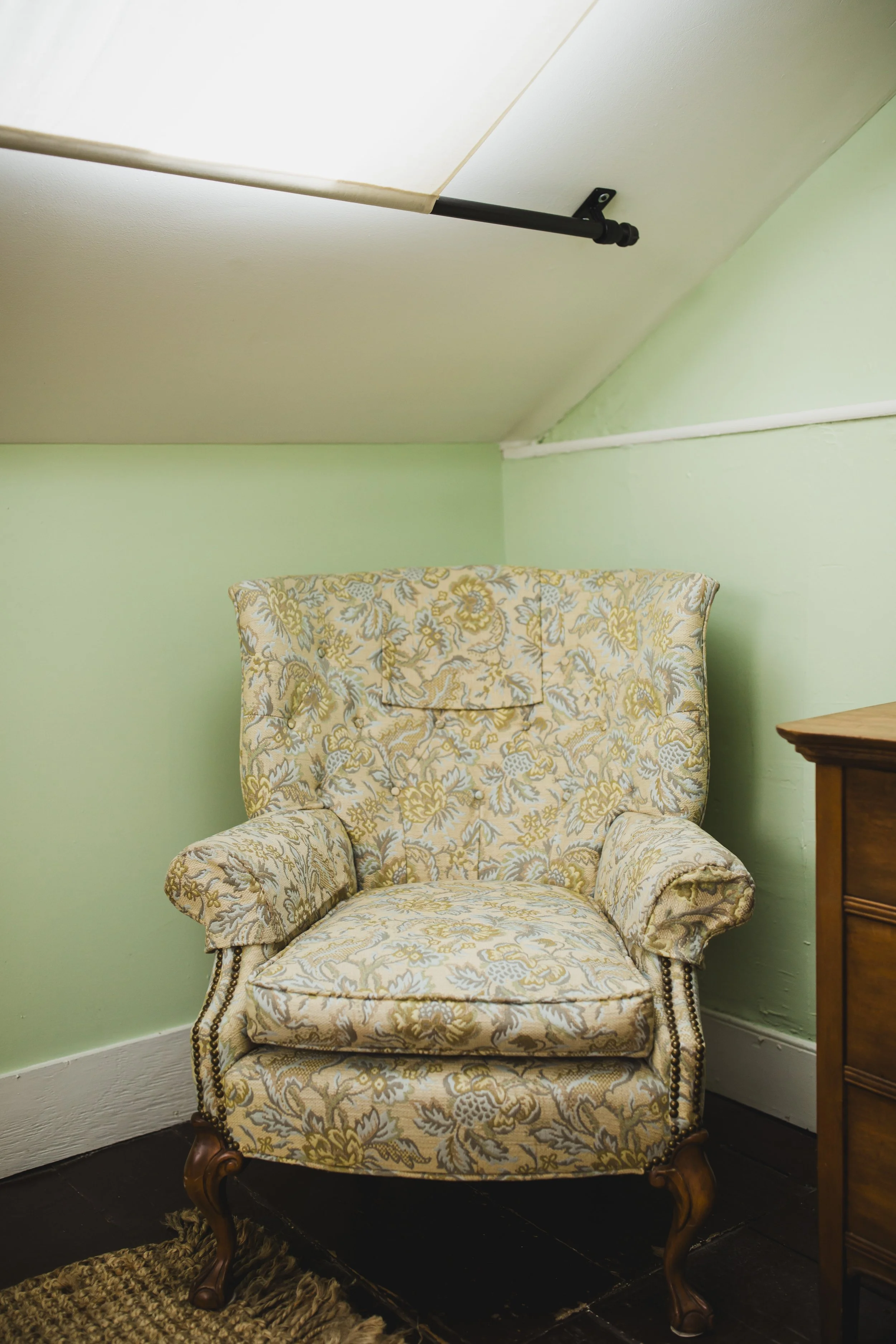 An upholstered armchair with a floral pattern, wooden legs, and a high backrest, placed in a room with green walls. There is a wooden piece of furniture on the right and a fringed rug on the dark floor. A portion of the ceiling with a ceiling mount a