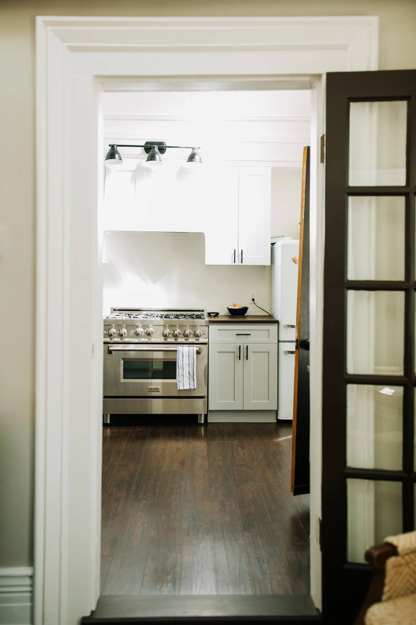 View through a doorway into a modern kitchen with white cabinets, a stainless steel stove, a black countertop, and a small black bowl on the counter. There is a wooden floor and a white ceiling with three black track lights.