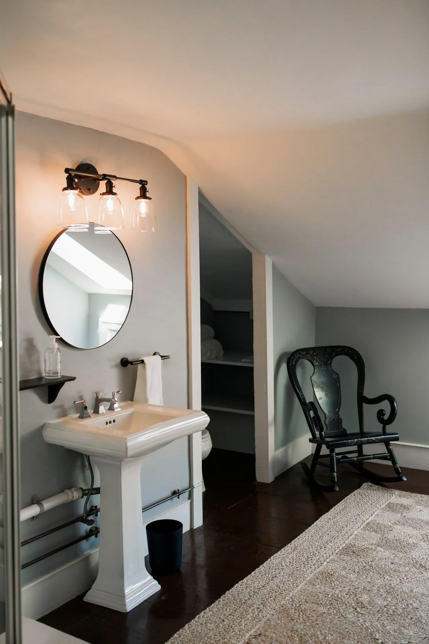 Bathroom with a white pedestal sink, oval mirror, black wall-mounted light fixture, black chair, and a beige rug.