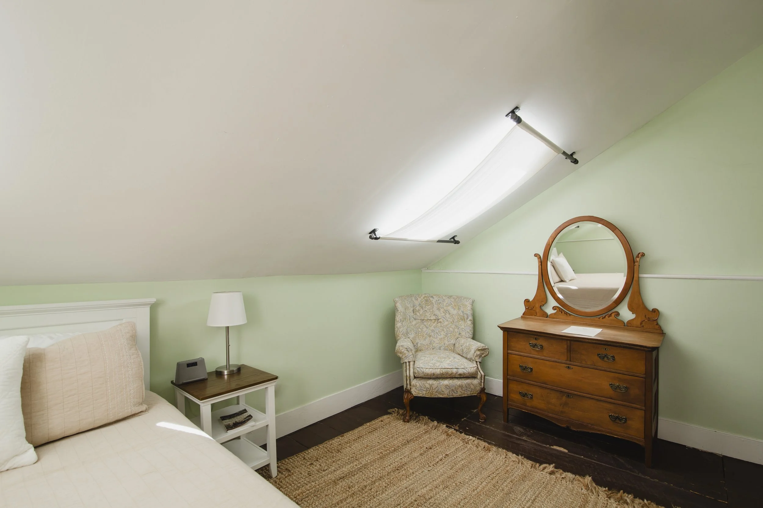 A bedroom with a slanted ceiling, green walls, a bed, a nightstand with a lamp and phone, an upholstered armchair, a wooden dresser with a mirror, and a star-shaped ceiling light fixture.
