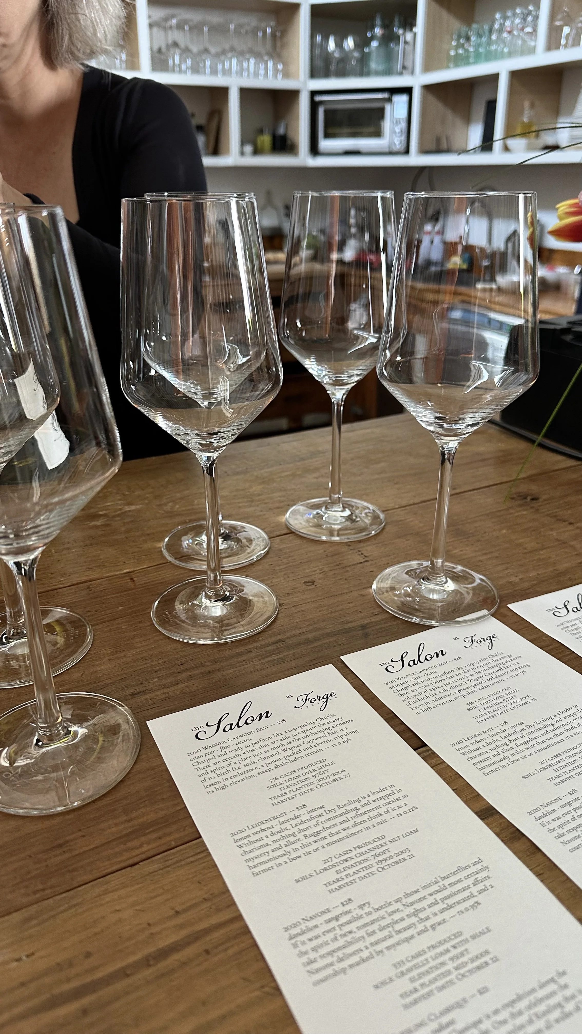 Multiple empty wine glasses on a wooden table with menus, in a cozy setting with shelves of glassware in the tasting room at Sheldrake Point Winery.