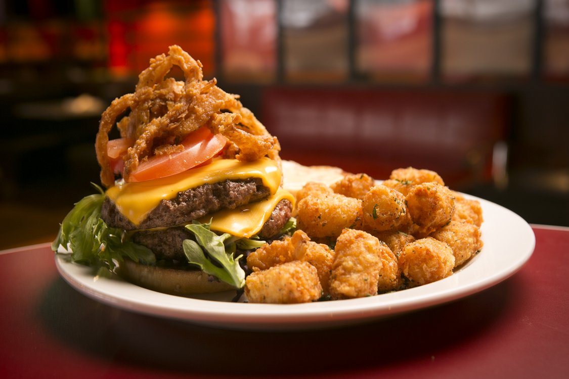 A plate of food at Atlas Bowl with a cheeseburger layered with lettuce, tomato, cheddar cheese, beef patty, and fried onions, accompanied by a side of tater tots.