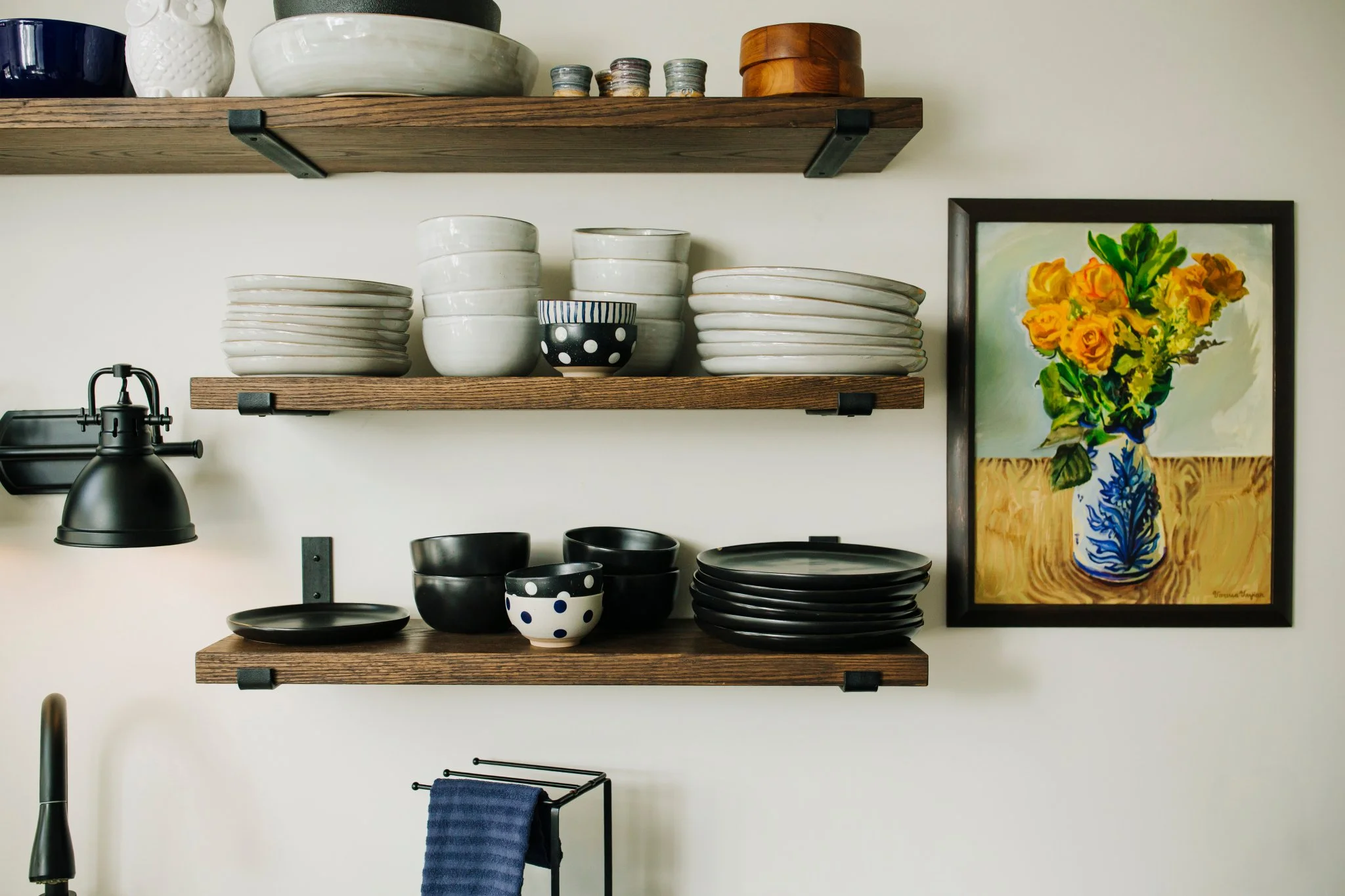 Open kitchen shelves displaying white and black dishware, a colorful framed floral painting on the wall, and a black wall-mounted lamp.