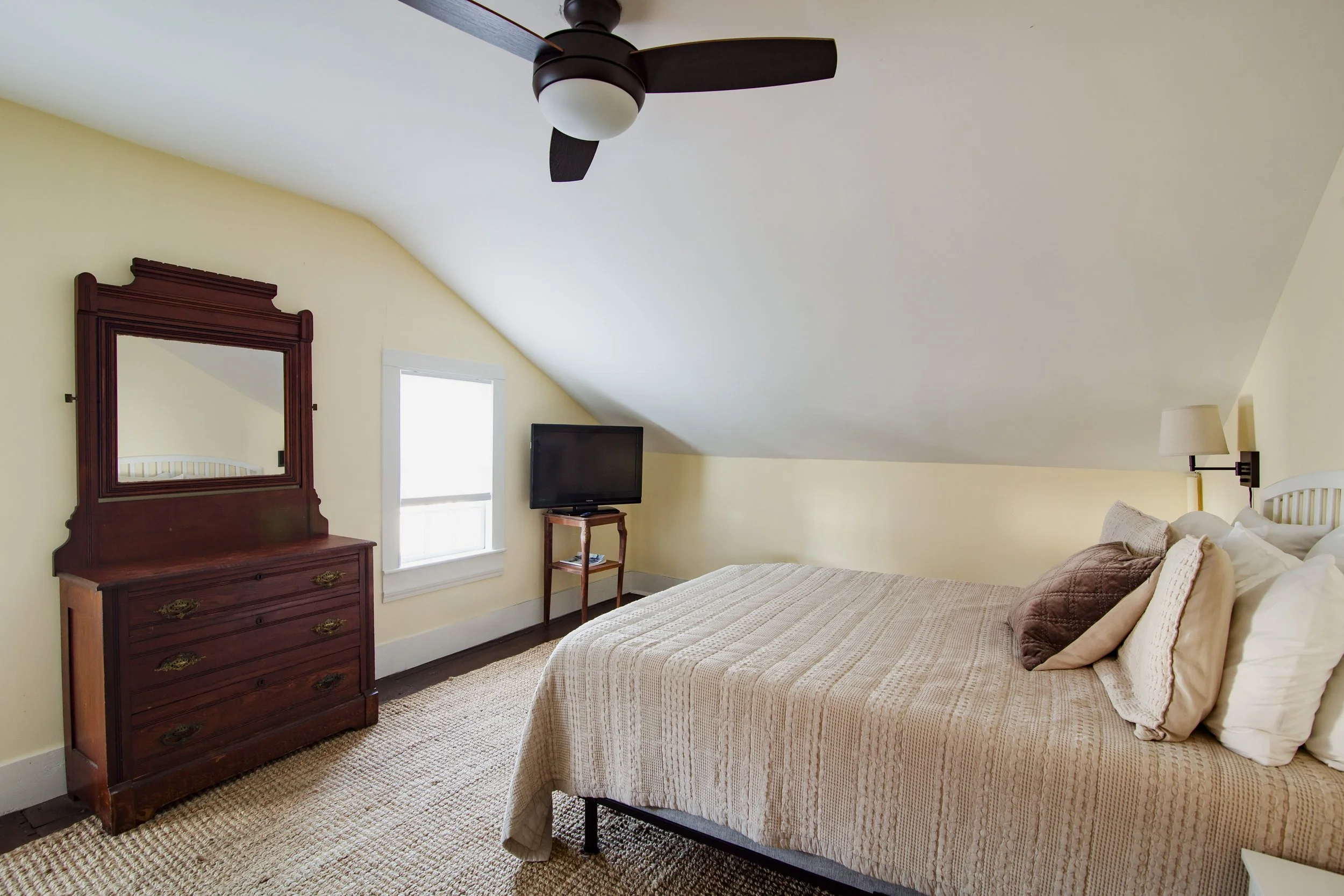 A bedroom with a sloped ceiling, a bed with beige striped bedding and pillows, a wall mirror with a wooden frame, a window, a small wooden table with a TV, a beige rug, and a ceiling fan.