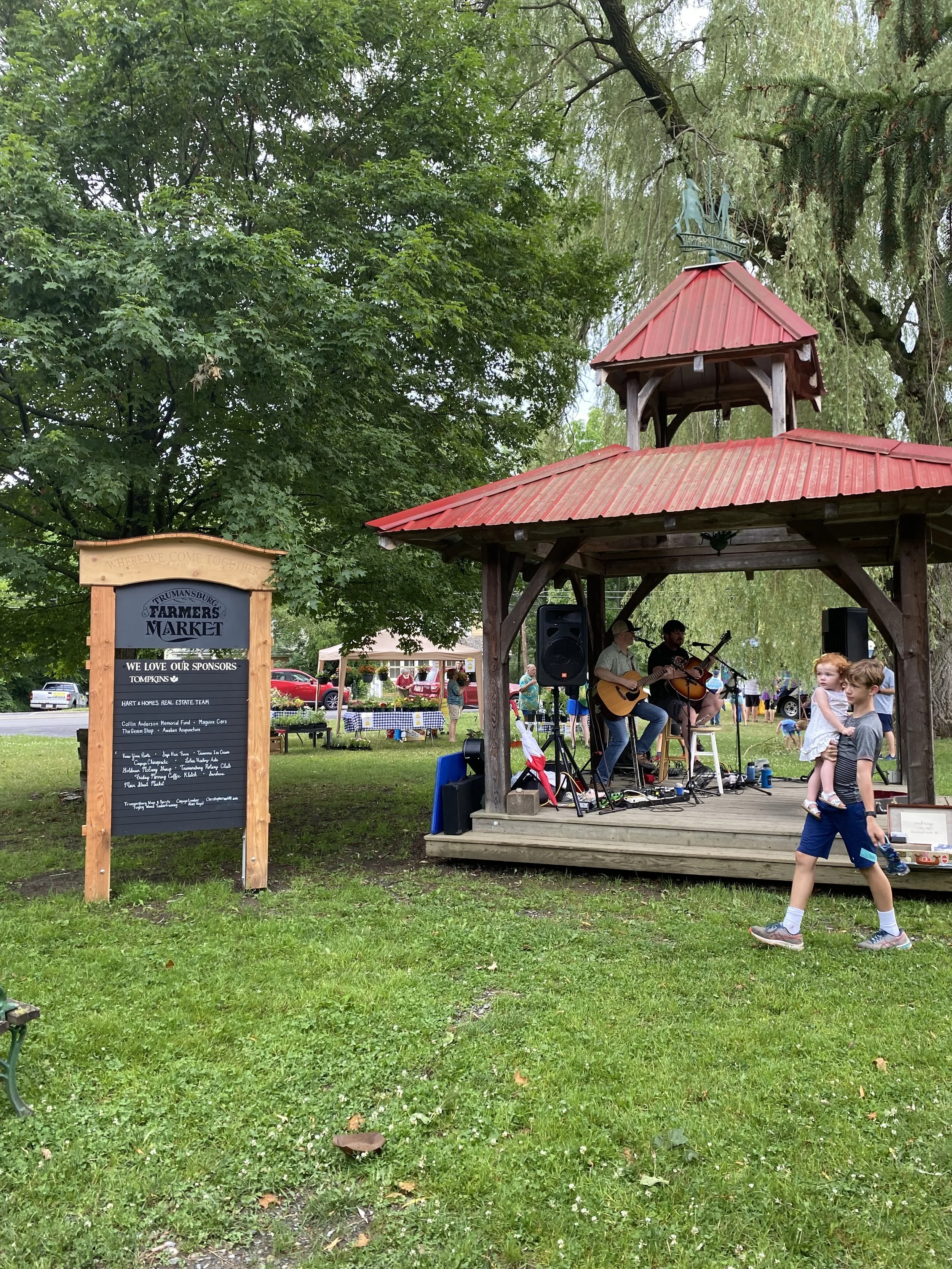 A small outdoor stage with musicians playing guitars, surrounded by greenery and people, at the Trumansburg Farmers Market farmers.