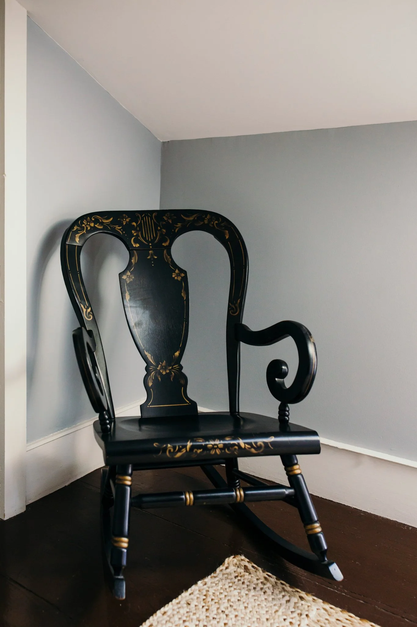 Black wooden rocking chair with gold floral designs, placed in a corner next to a beige wall with white trim and dark hardwood floor. A small beige woven rug is partially visible in front of the chair.