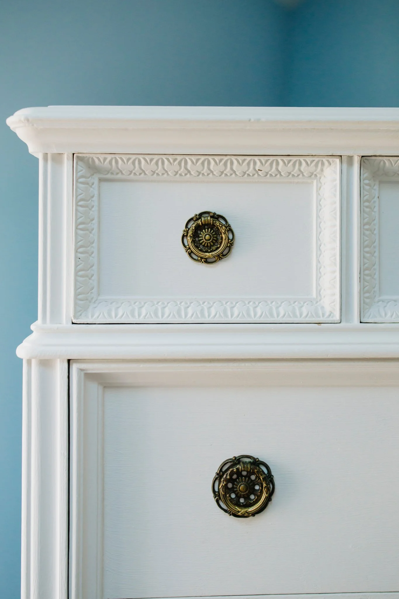 Close-up of a white wooden dresser with decorative carved details and ornate brass handles, set against a blue wall.