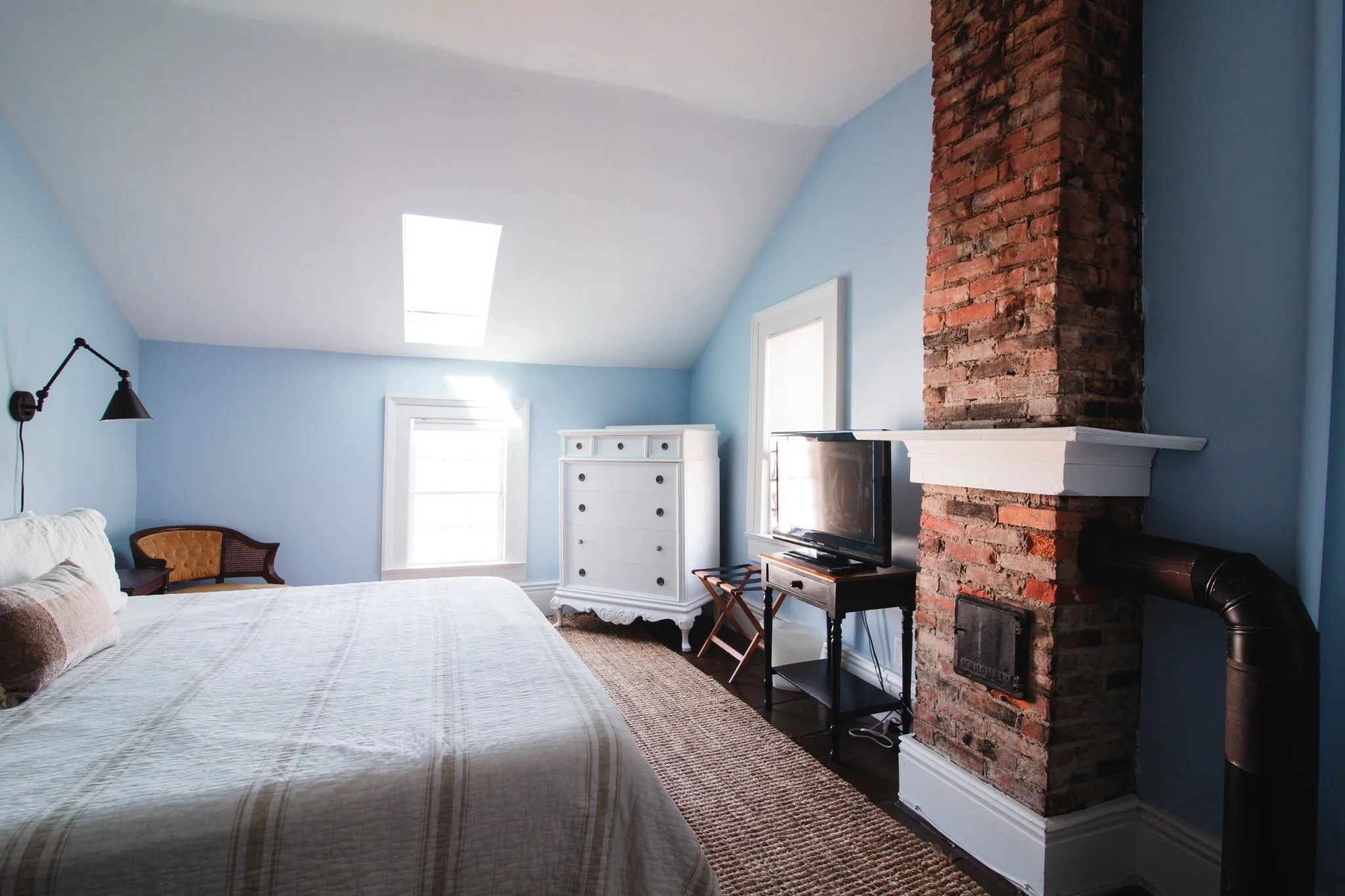 A bedroom with light blue walls and a sloped ceiling with a skylight. There is a white bed with beige and brown pillows, a black wall-mounted lamp, a yellow cushioned chair, a white dresser, a window, and a brick chimney with a white mantel and a small black TV stand with a television. A flat-screen TV is on the stand, and a black pipe extends from the chimney.