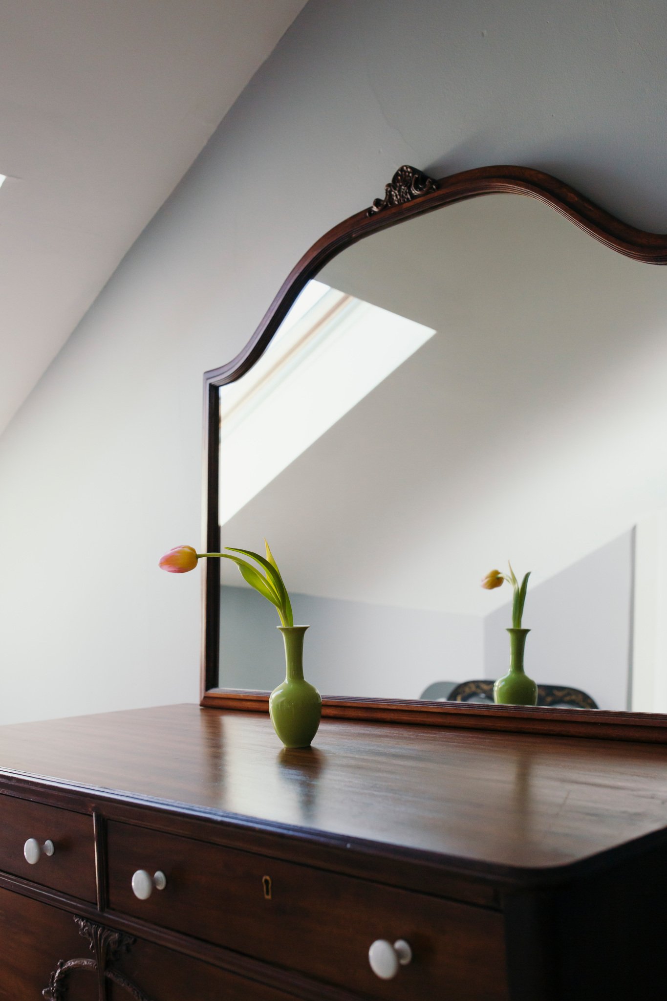 A wooden dresser with white knobs, a mirror with a wooden frame, and two green vases with pink tulips on top of the dresser in front of the mirror.