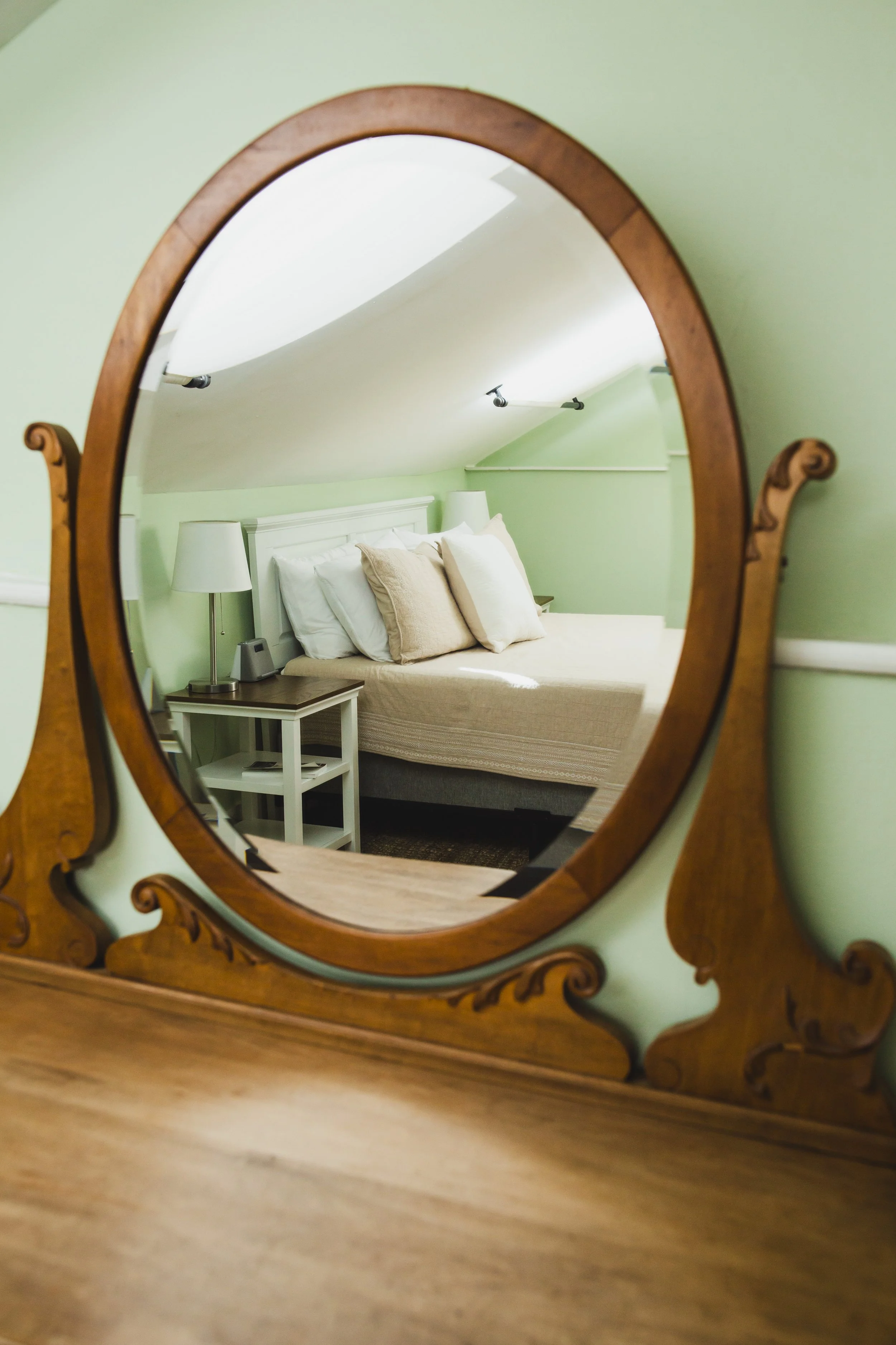 A bedroom seen through an ornate wooden framed mirror. The room has a white bed with beige pillows, a white side table with a lamp and a small speaker, green walls, and ceiling track lighting.