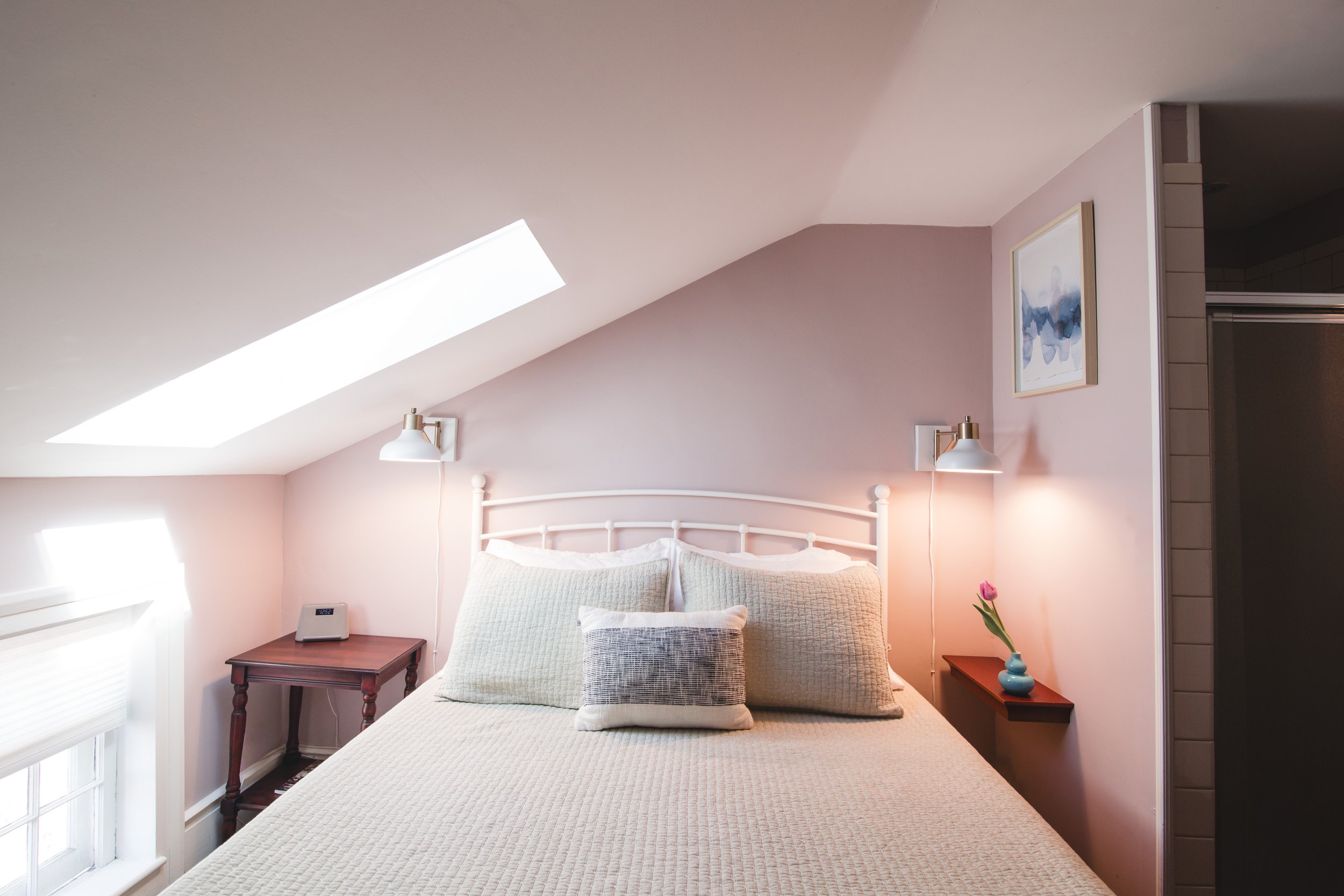 A cozy bedroom with a sloped ceiling featuring a skylight window. The bed has white bedding, pillows, and a woven blanket. There are two wall-mounted lamps and a framed abstract blue and white painting above the bed. A small wooden side table holds a