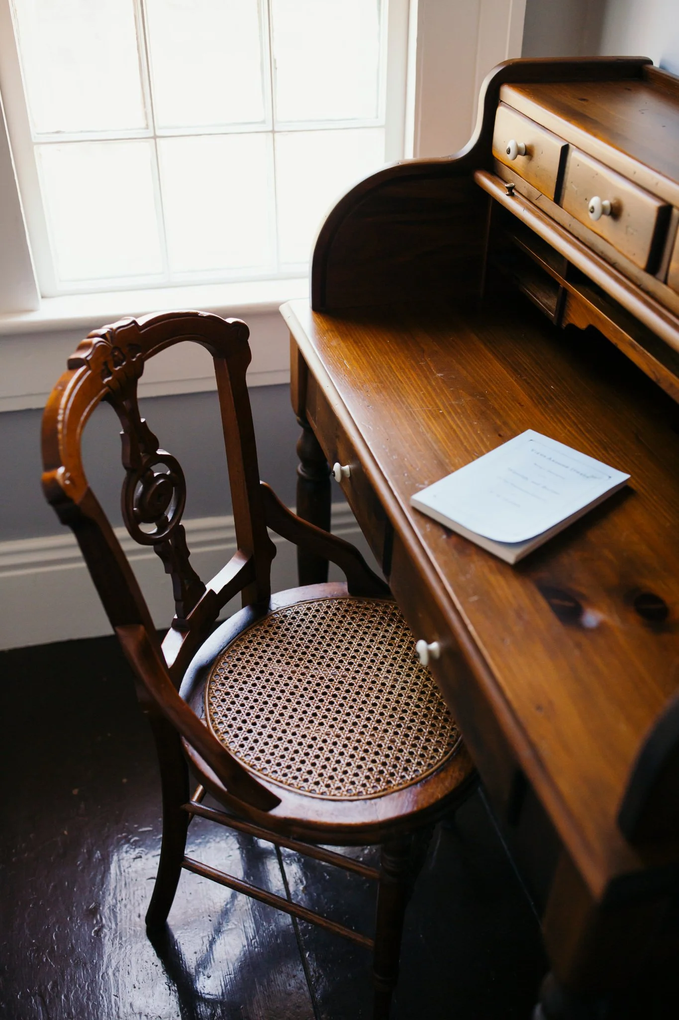 A wooden desk and a wooden chair with a woven wicker seat are positioned in front of a window with white panes. There is a notebook or paper on the desk.