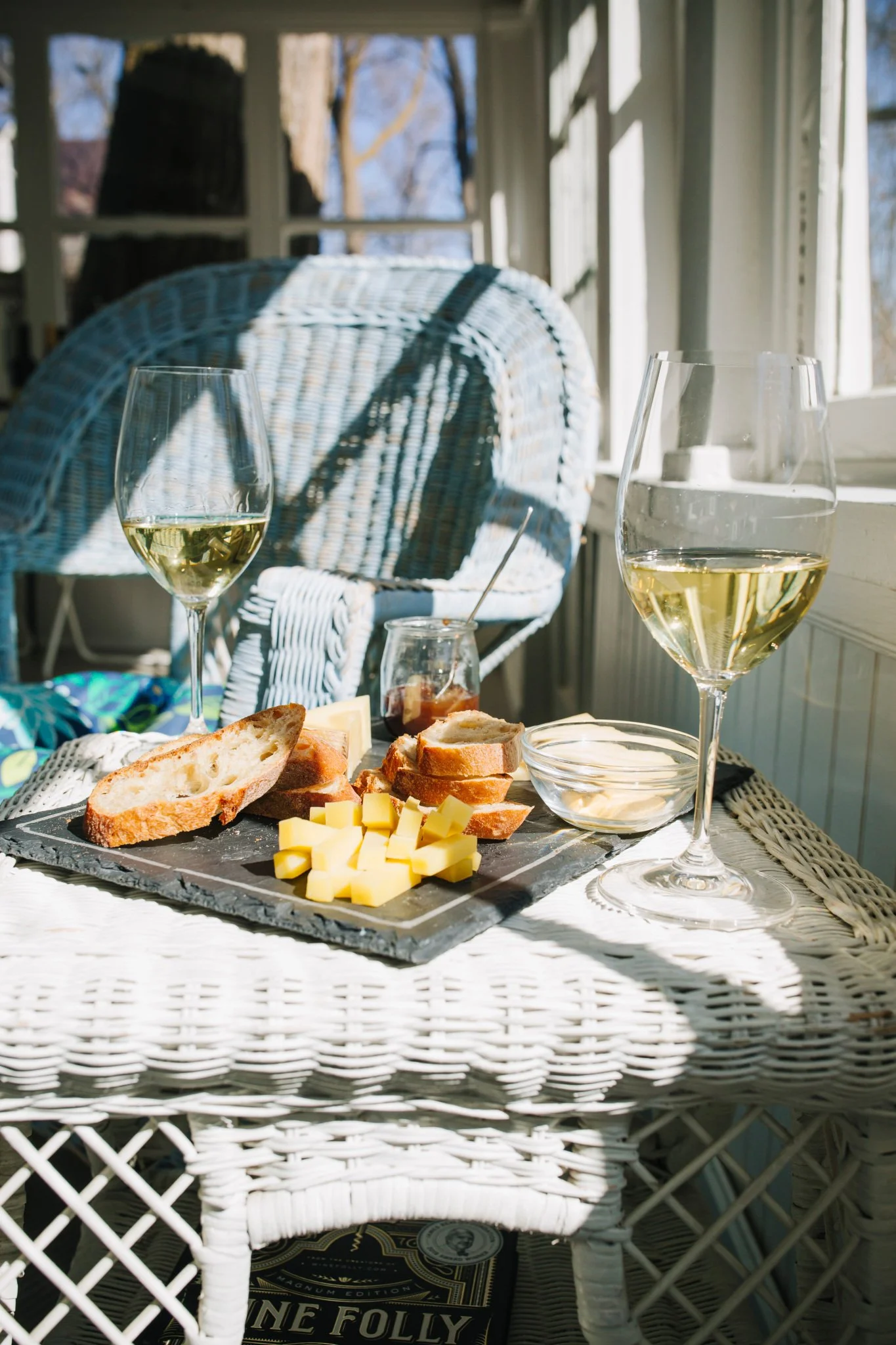 A table set with wine glasses, a cheese platter, bread, and condiments in a sunlit porch with wicker furniture and a view of trees outside.