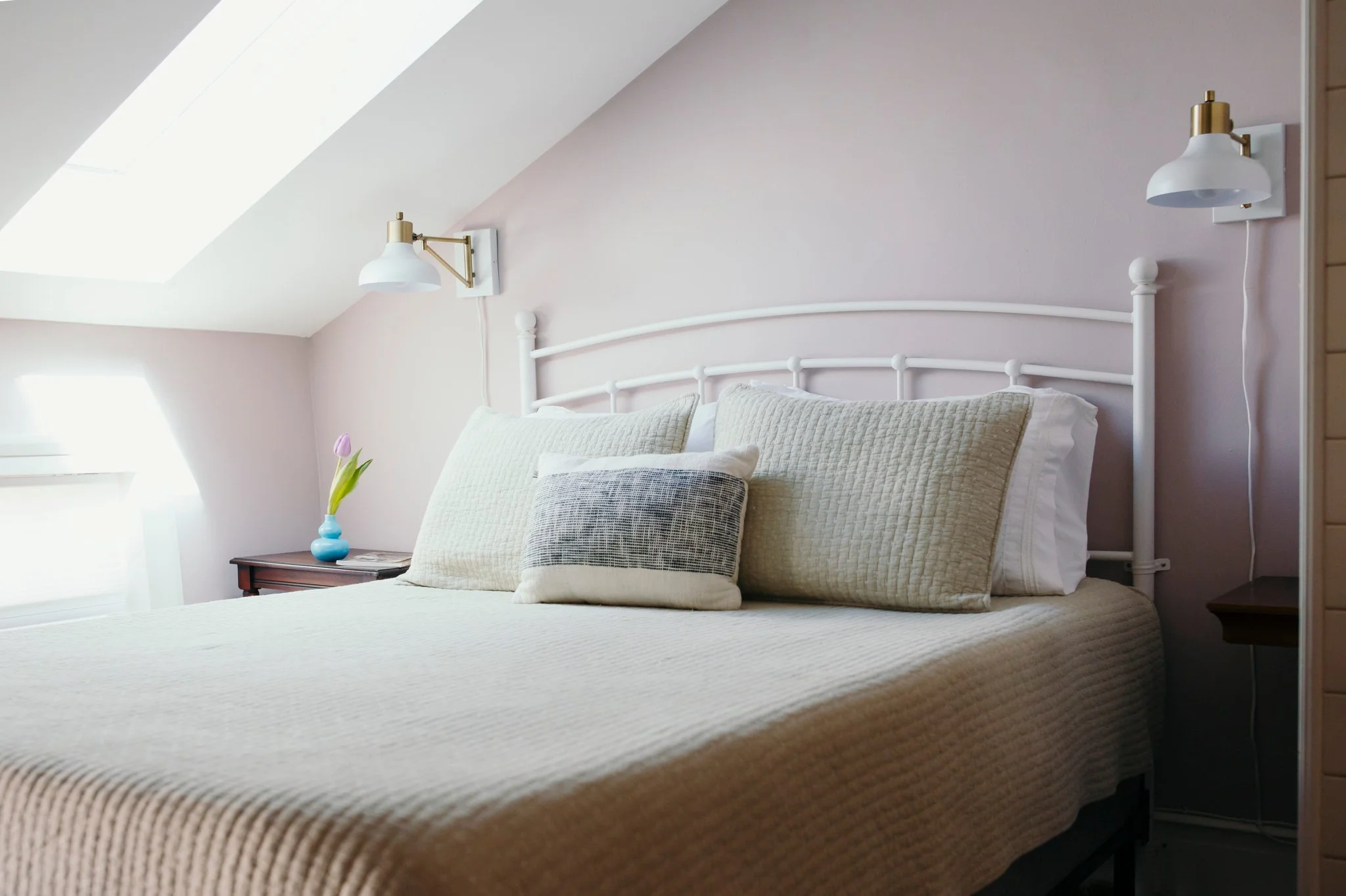 A neatly made bed with multiple pillows topped with a beige quilt, set against a soft pink wall. On each side of the bed, there are white wall-mounted lamps with brass accents. A small wooden nightstand with a blue vase of pink tulips is beside the bed. Natural light enters through a skylight window on the sloped ceiling.