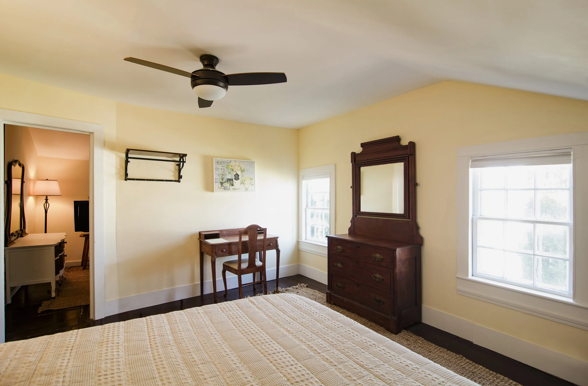 A bedroom with light yellow walls, a dark wood dresser with a mirror, two large windows, a small wooden desk and chair, and a ceiling fan with a light. An adjacent room is visible through an open doorway, showing a lamp and mirror.