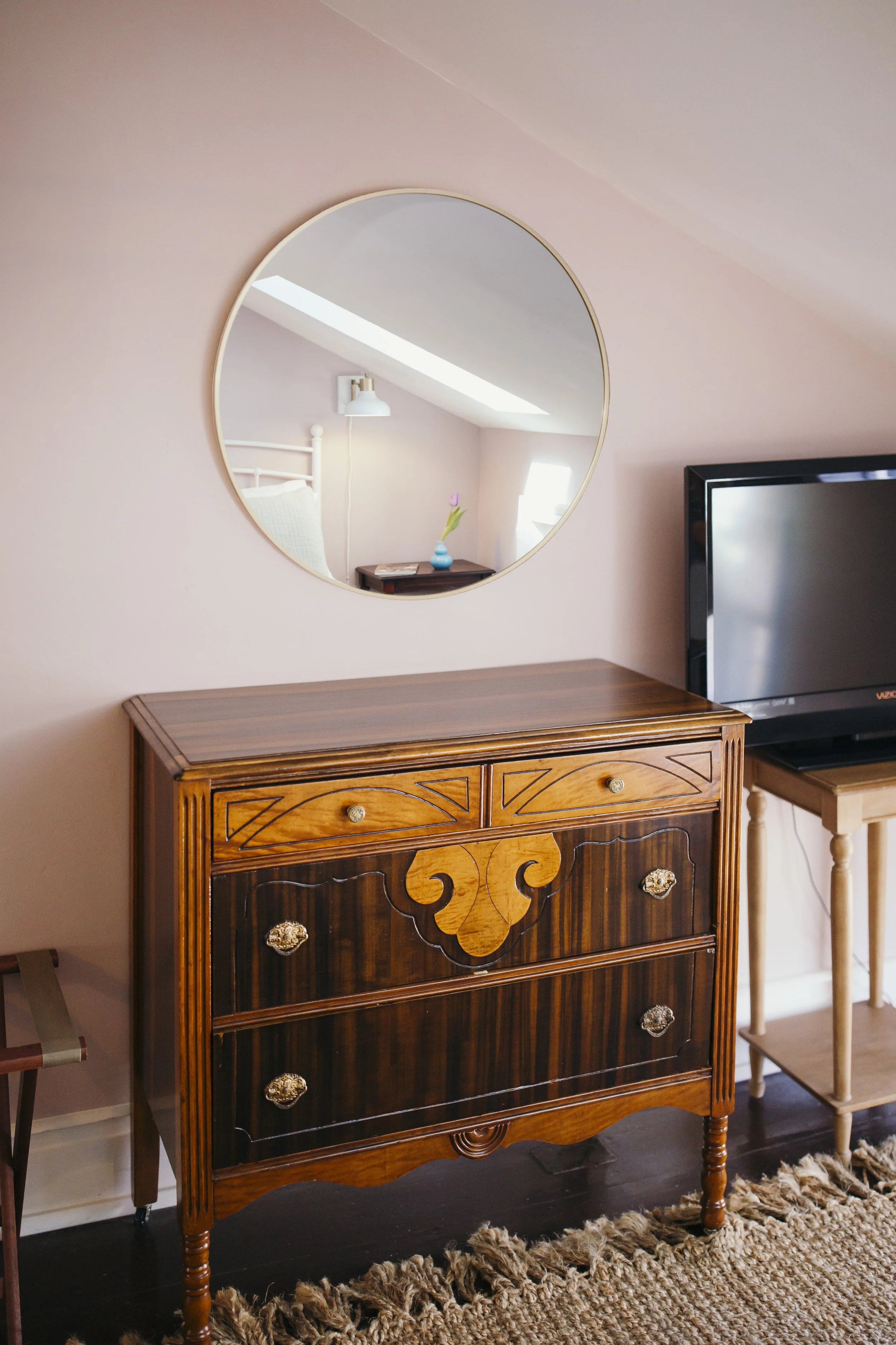 A wooden dresser with ornate inlay and brass handles, a round mirror on the wall above it, and a flat-screen TV to the right. There is a beige shaggy rug on dark wood flooring and a small chair to the left of the dresser.