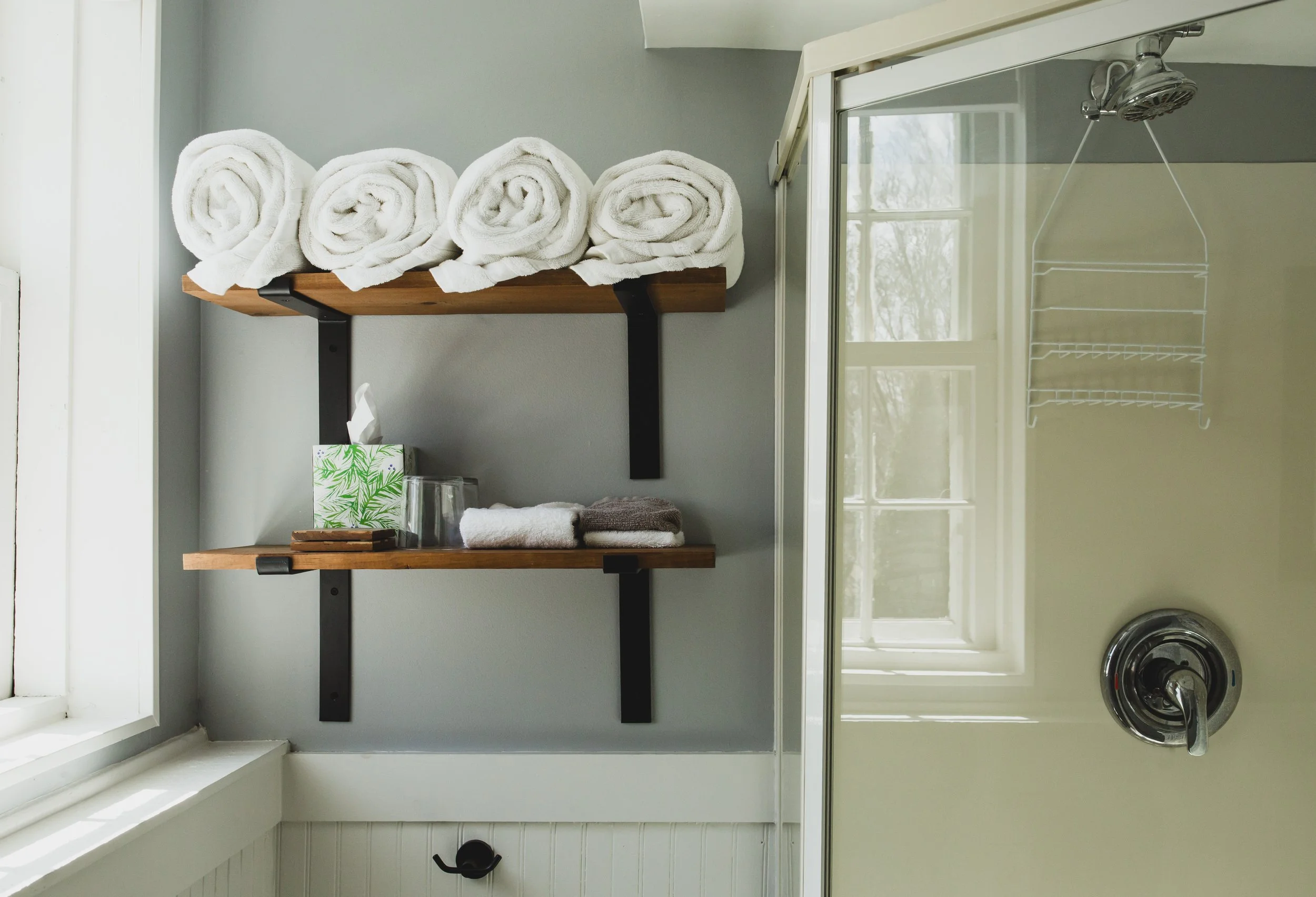 A bathroom shelf with rolled white towels, a tissue box with green leaves, drinking glasses, and some folded towels, next to a glass shower enclosure with a showerhead and a drain in the floor.
