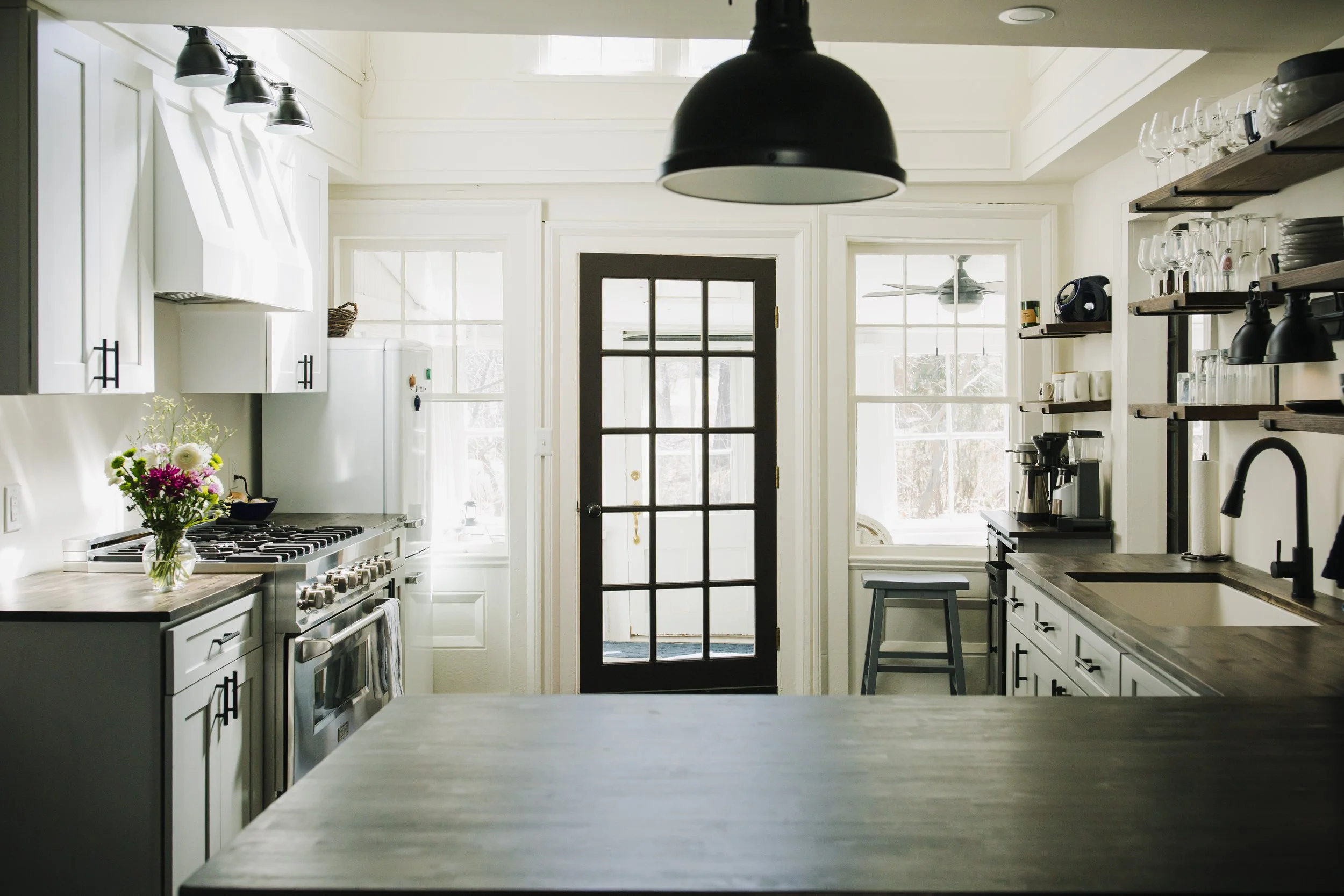 Bright kitchen with white cabinets, a black door with glass panes, and open shelves with dishes and glasses. A bouquet of flowers is on the counter, with a stove and coffee maker visible. Large windows and a ceiling fan provide natural light.