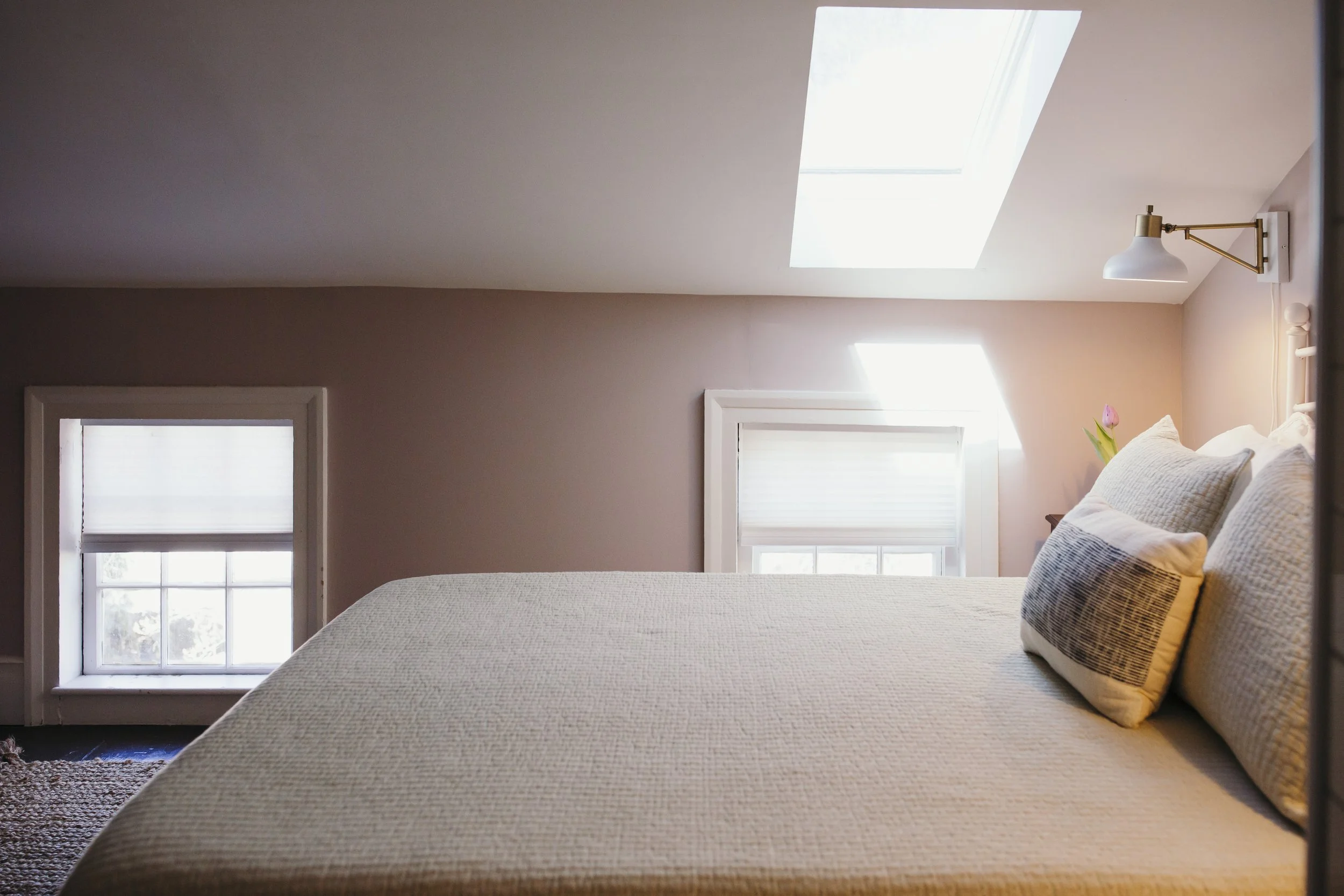 A bedroom with a bed, pillows, and a window with sunlight coming through, featuring beige walls and a skylight.