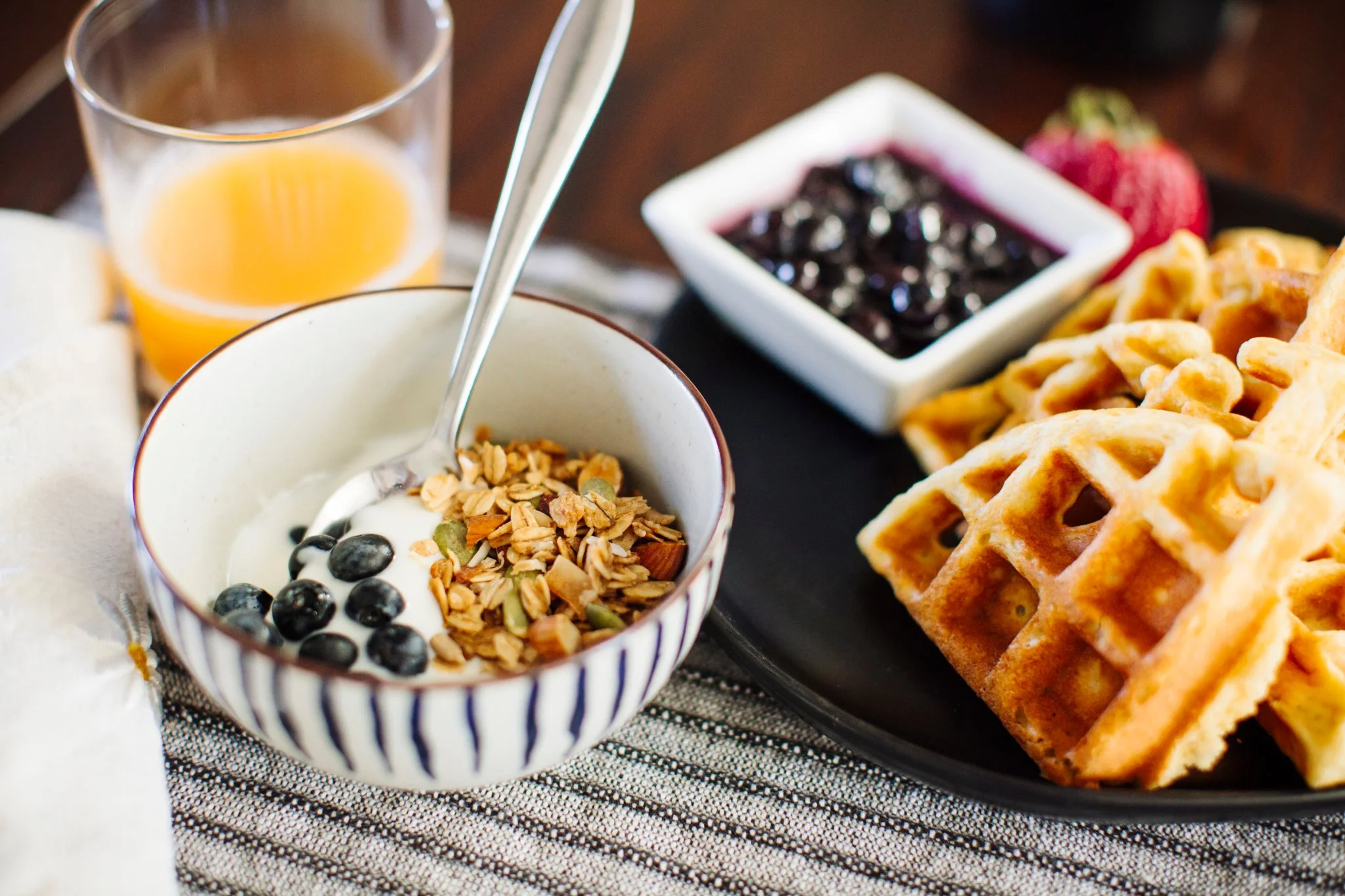 Close-up of a homemade breakfast spread including a bowl of yogurt with granola and local blueberries, a glass of orange juice, a square dish of blueberry jam or syrup, and two waffles on a black plate.