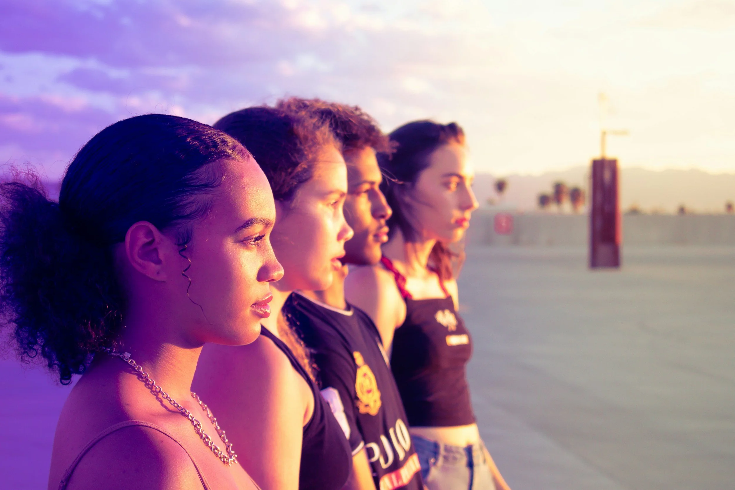 Four women standing side by side outdoors during sunset, facing left with serious expressions.