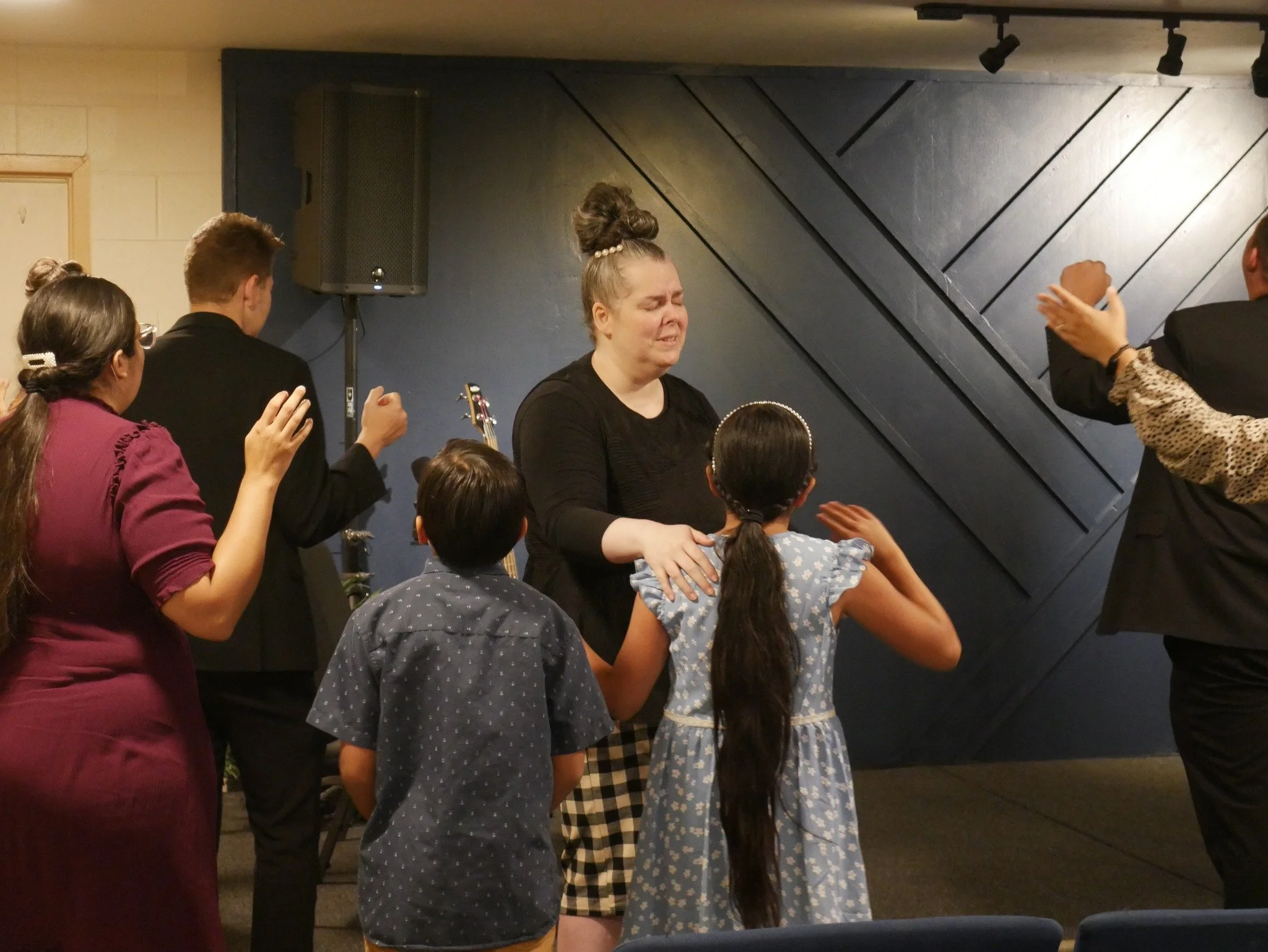 A group of people, including a woman with closed eyes and tears, appears to be praying or engaging in a spiritual moment in a church or religious setting. A woman with long hair and two children are at the center, with others surrounding them, some with hands raised in prayer.