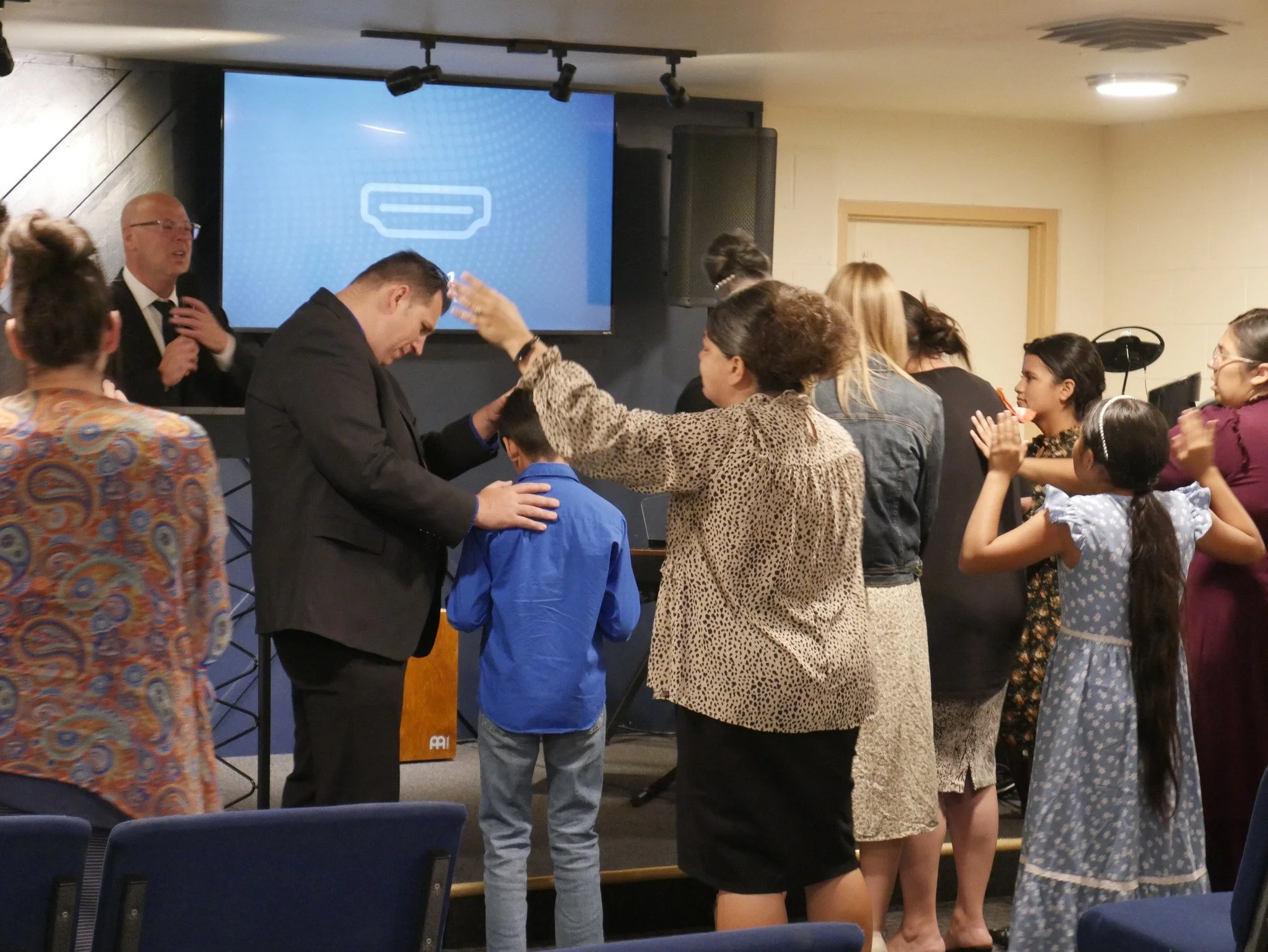 People participating in a religious ceremony or service, with a man receiving a blessing or prayer from a woman, while others are clapping and watching in the background, inside a church or community center.
