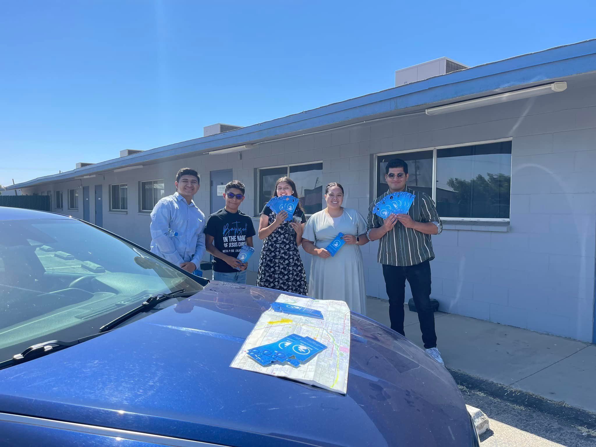 Group of five young people standing outside a building, holding blue pamphlets, with a car and a map on its hood in the foreground.