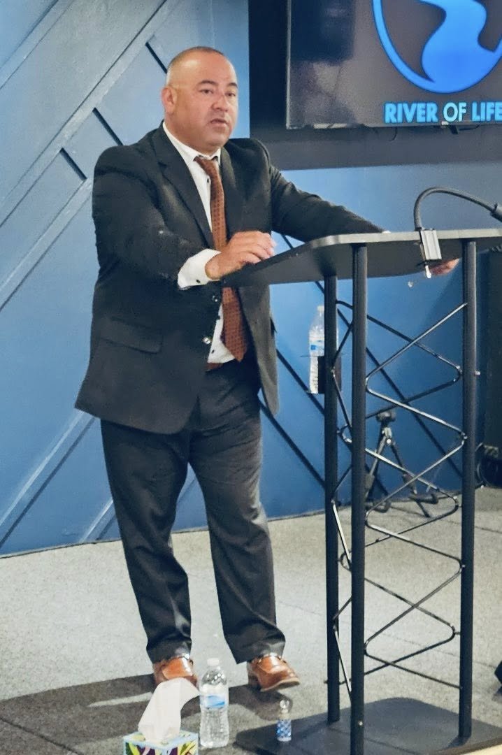 A man in a suit and brown tie standing behind a podium, speaking at a conference or presentation in front of a blue and black background with a logo that reads 'River of Life'.