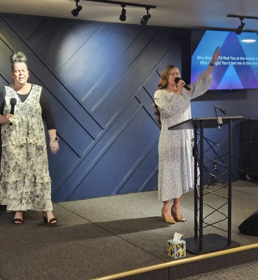 Two women are singing at a church or worship service, one holding a microphone and raising her hand, with a screen displaying lyrics behind them.