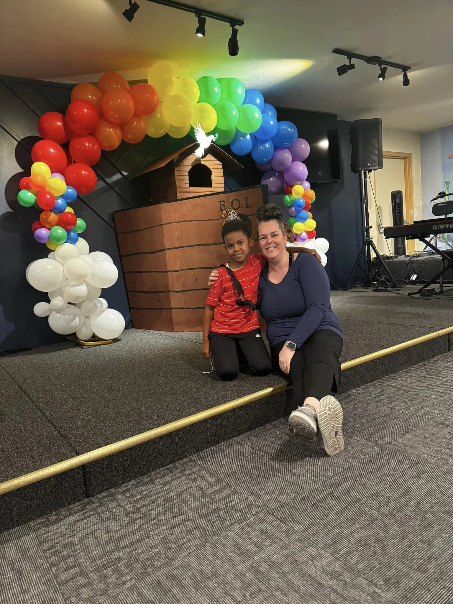 A woman and a girl sitting on a stage, smiling at the camera. The stage is decorated with colorful rainbow balloons and a small nativity scene with a dove above it.