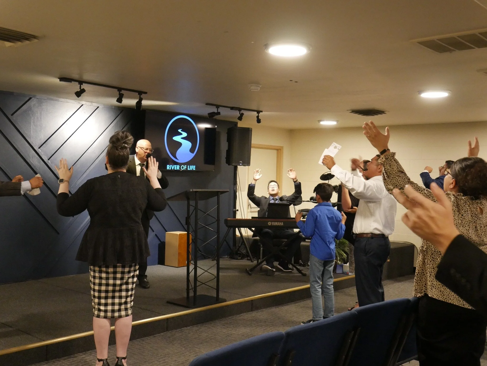 People worshiping in a church or religious setting, with a man speaking at the front, a woman with arms raised, a woman playing a keyboard, and others raising their hands or holding papers.
