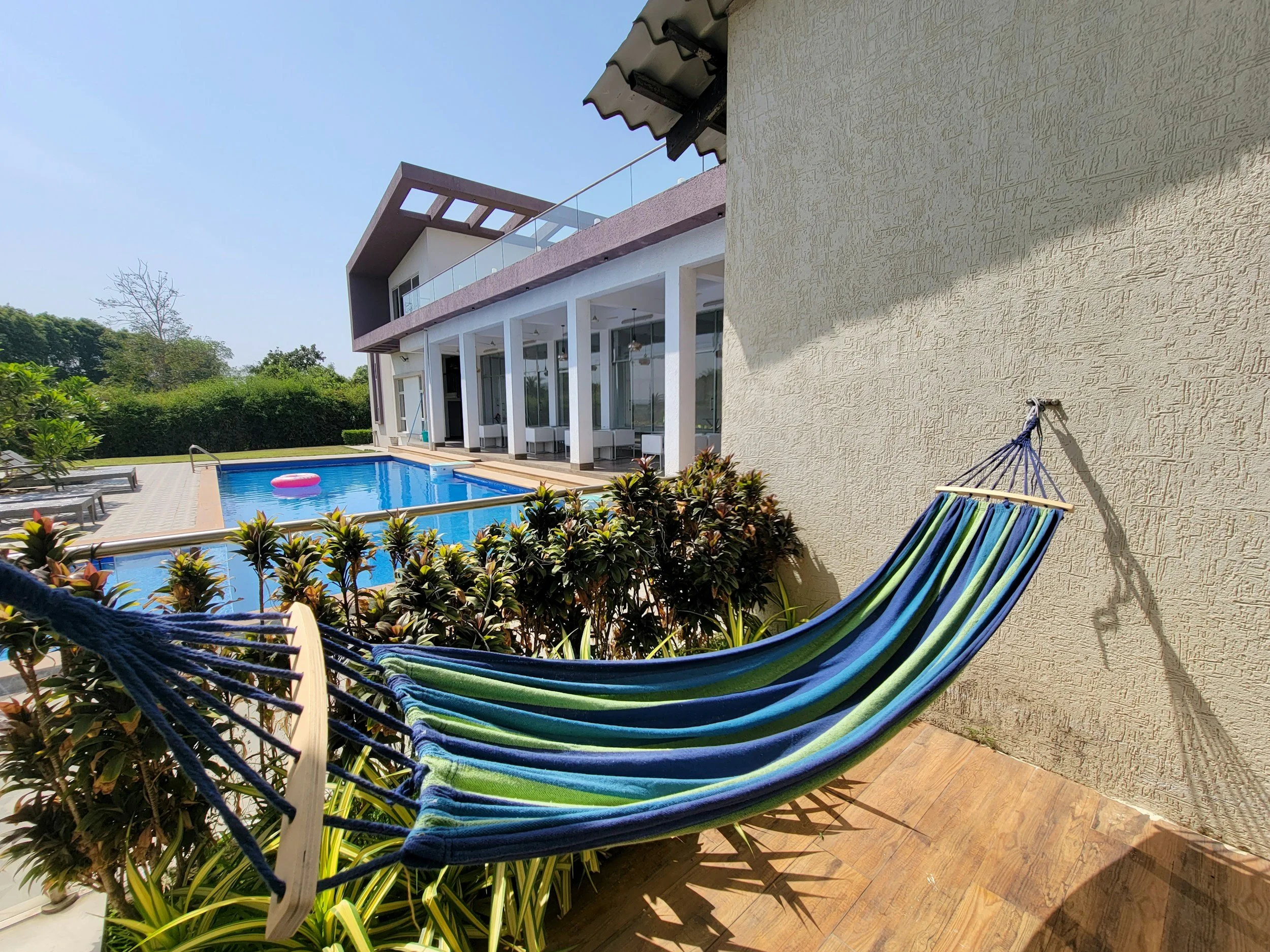 Colorful striped hammock on a patio near a swimming pool with lounge chairs and a modern house in the background.