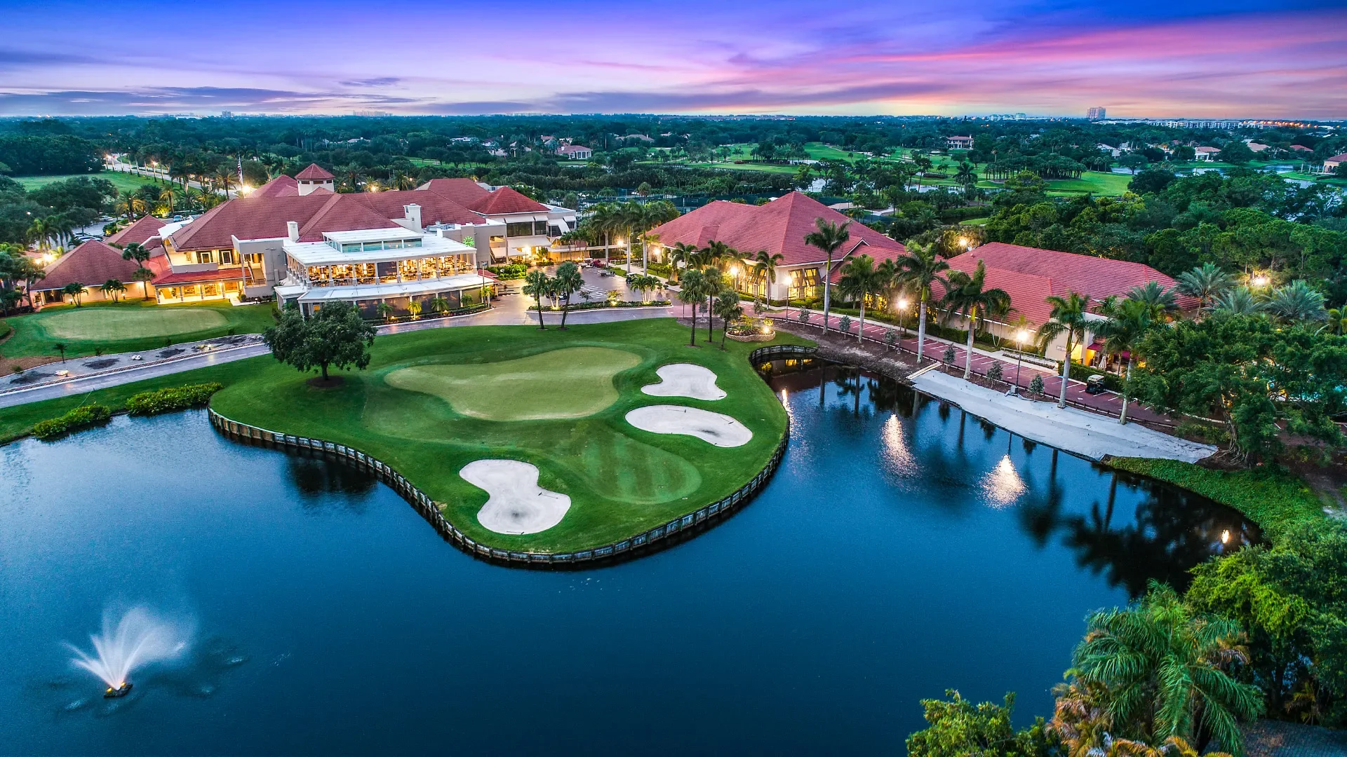 Aerial view of a golf course with a water hazard, surrounded by luxury buildings and lush greenery at sunset.