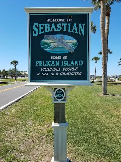 Welcome sign for Sebastian, Florida, featuring an illustration of a fish, with text welcoming visitors, mentioning Pelican Island, friendly people, and six old grouches, and displaying the establishment year 1924.