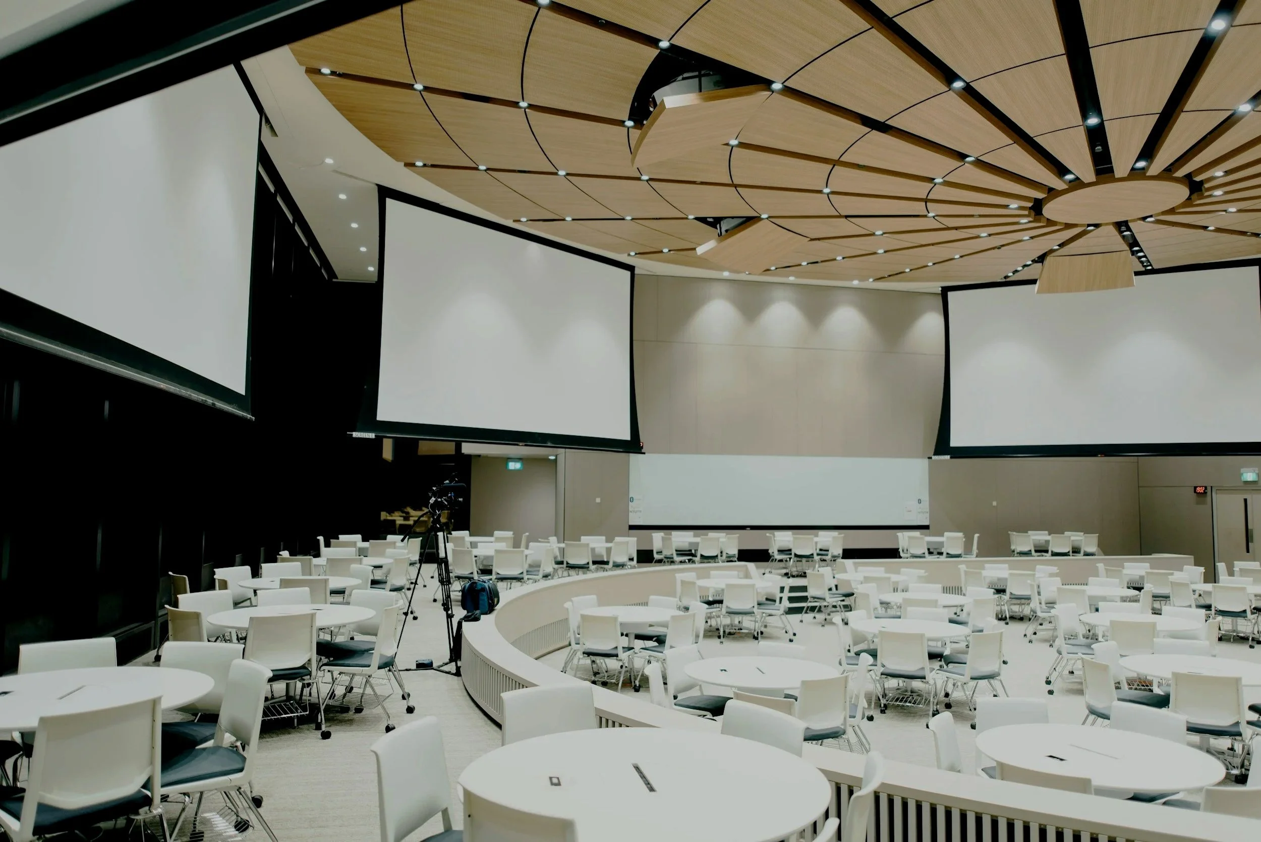 A modern conference room with three large projection screens on the walls, white tables and chairs arranged in a semi-circle, and a ceiling with wooden panels and integrated lighting.