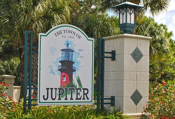 Sign for the town of Jupiter with a lighthouse illustration, surrounded by palm trees and plants.