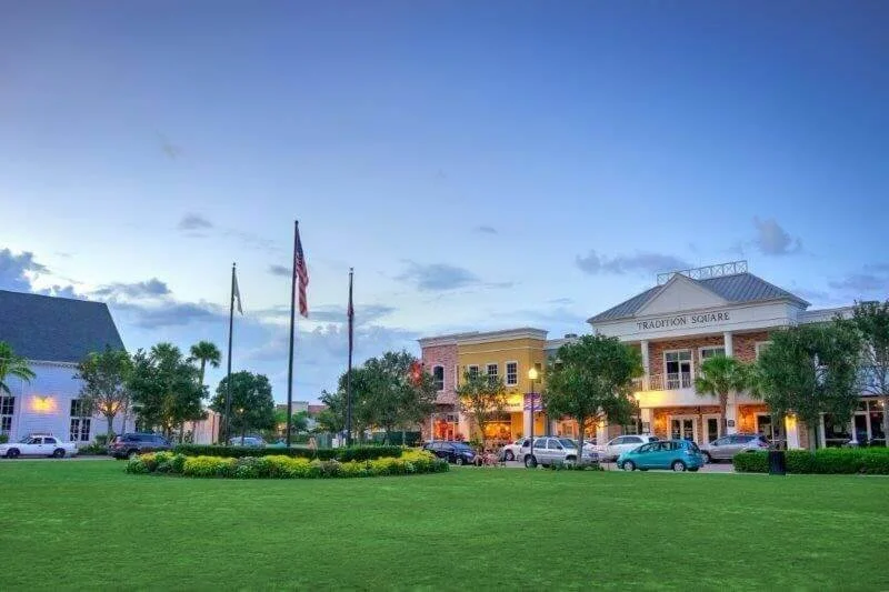 A shopping center called Tradition Square with parked cars, trees, and an open grassy area under a sky with some clouds.