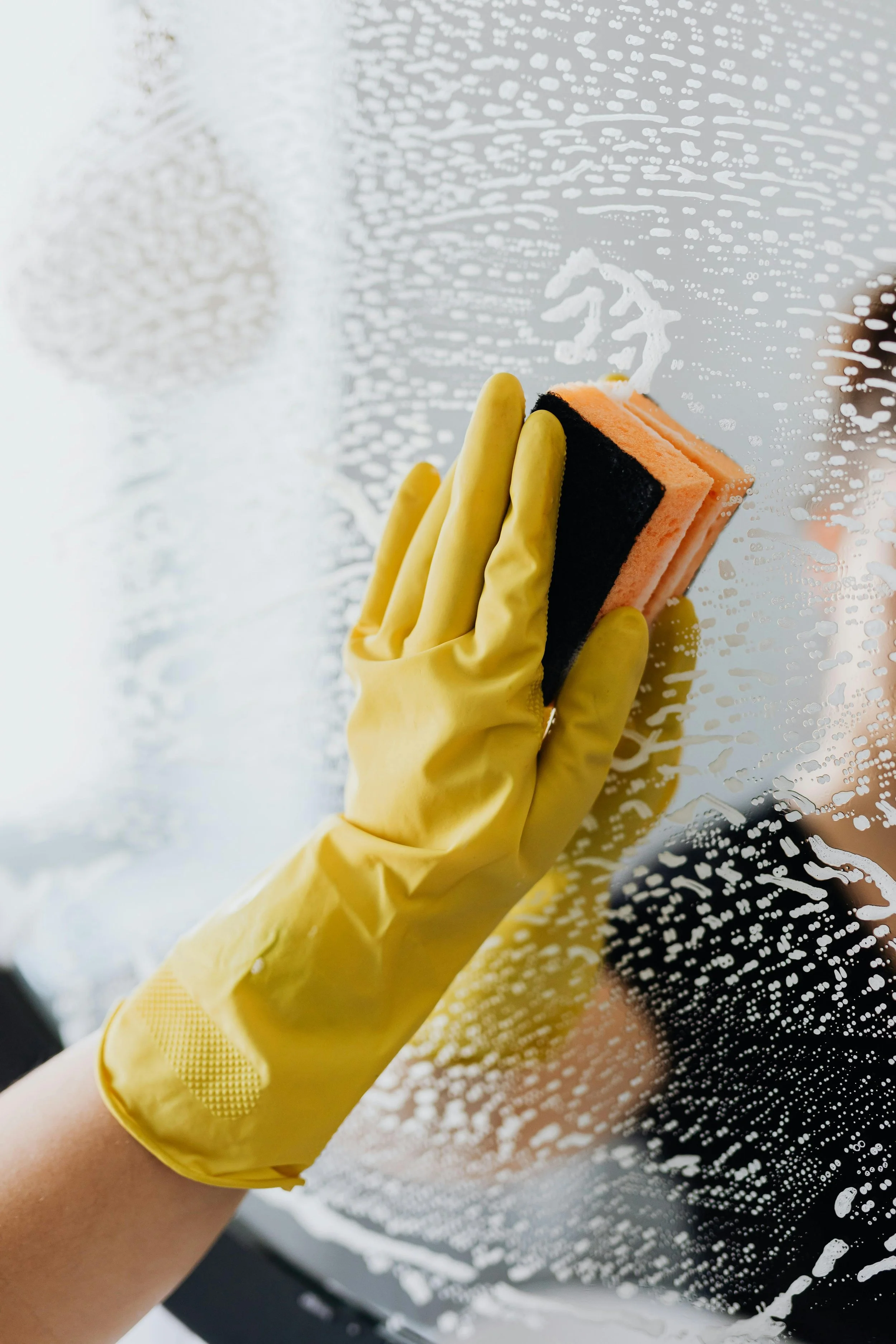A person wearing yellow rubber gloves, cleaning a window with a sponge and soap.
