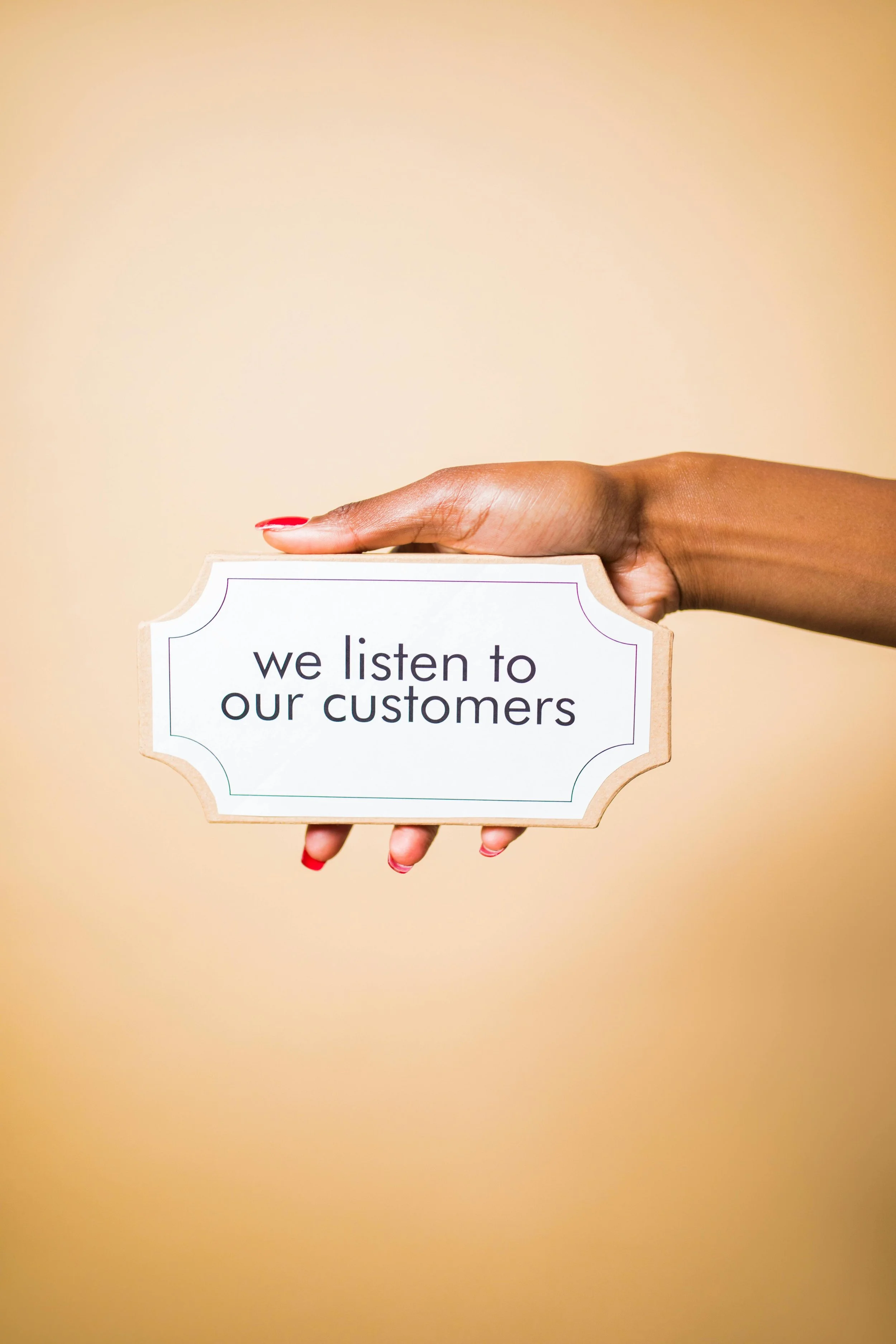 A person's hand with red painted nails holding a sign that says "we listen to our customers" against a beige background.