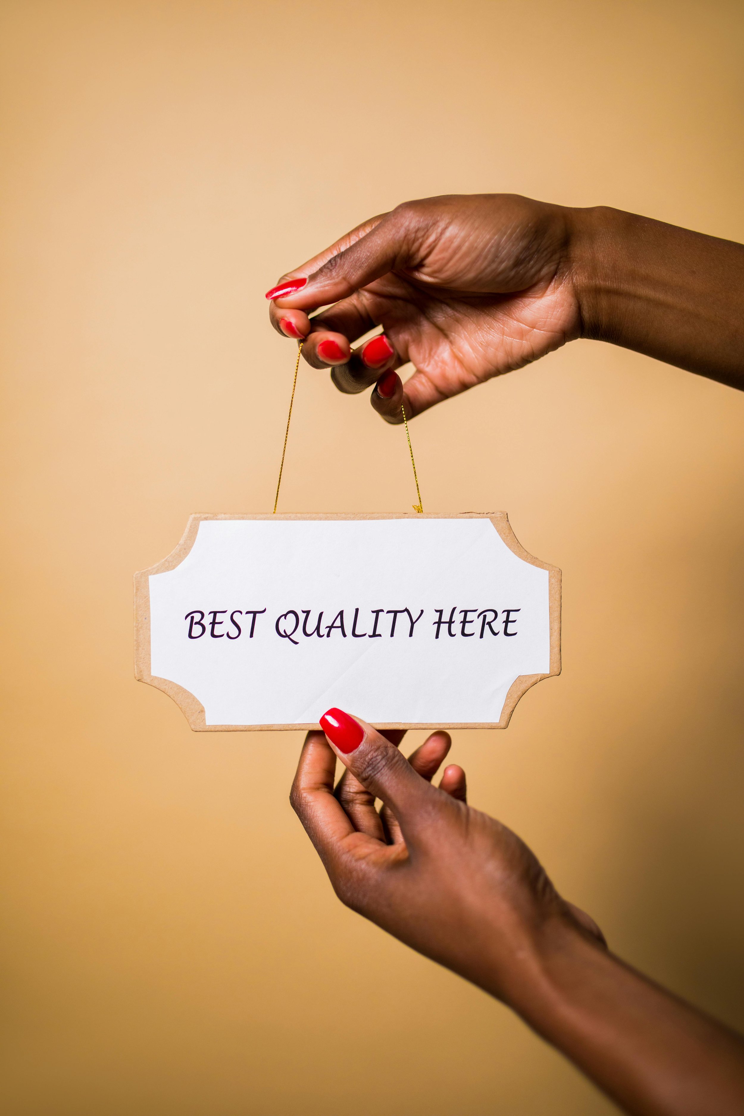 Two hands holding a sign with the words "Best Quality Here" written on it, against a beige background.