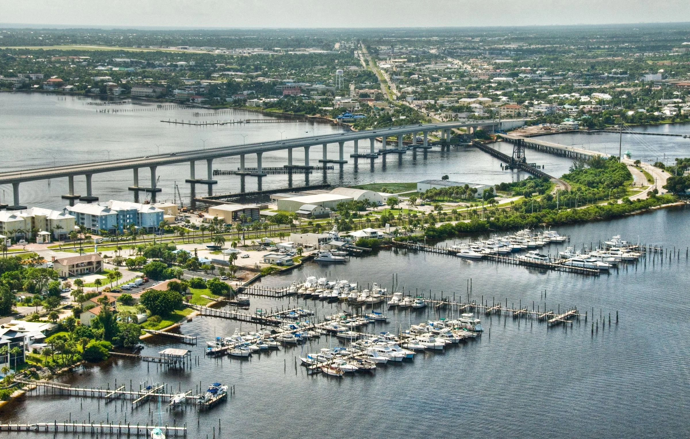 Aerial view of a marina with boats docked, a bridge crossing a river, and a cityscape in the background.