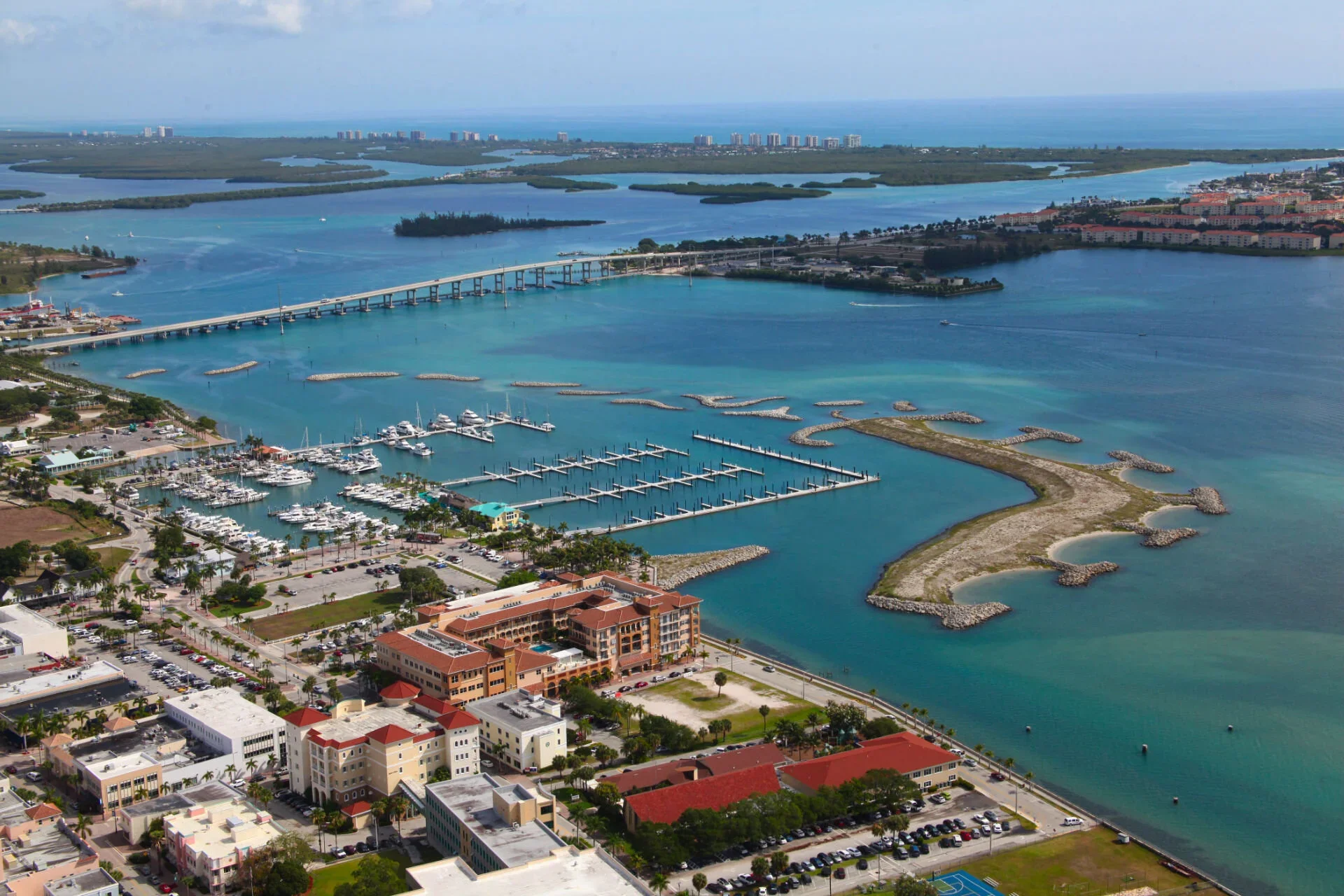 Aerial view of a marina with boats docked in the water, surrounded by buildings, roads, and a bridge over a body of water, with islands and a distant city skyline in the background.