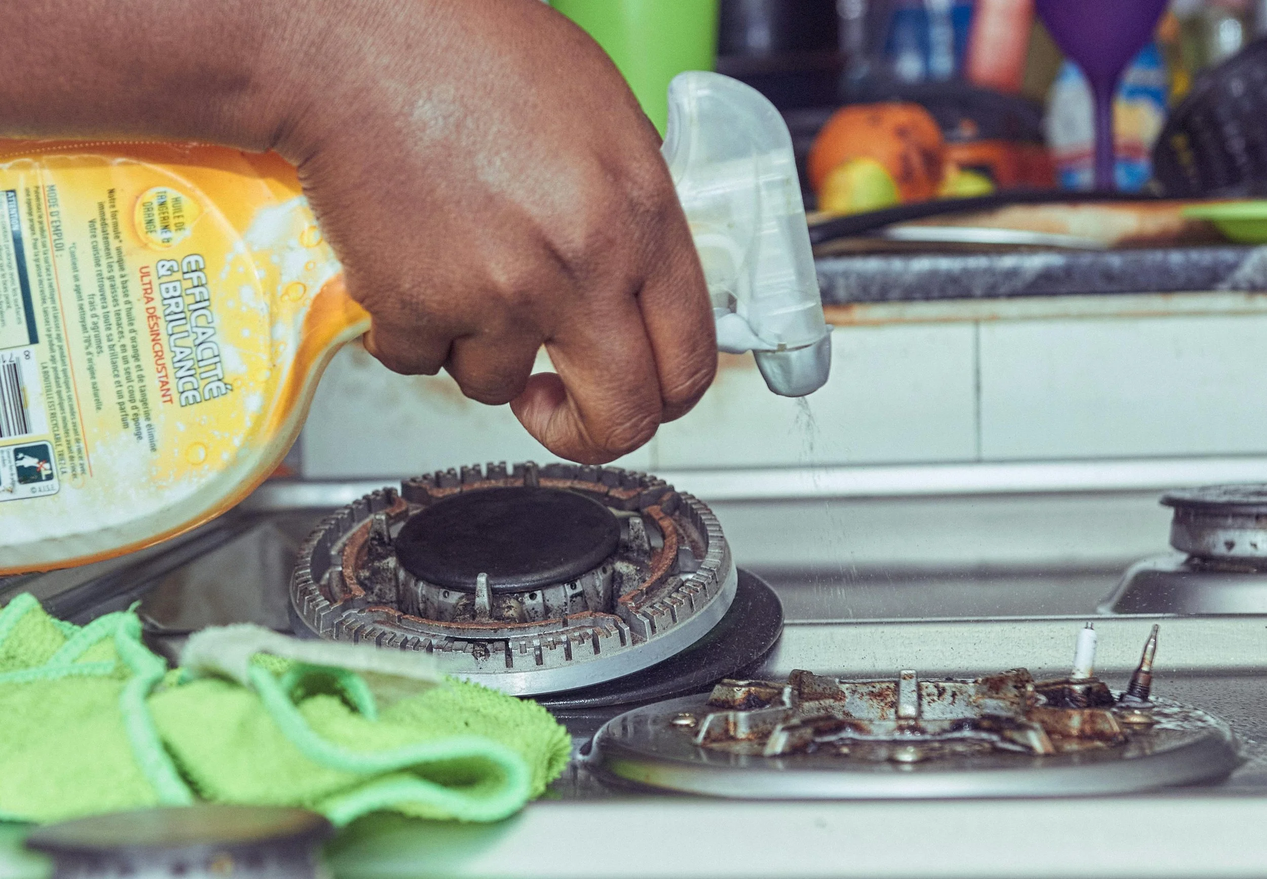 A person's hand sprays cleaning solution onto a stovetop burner. The stove has signs of rust and grime, with a green cloth nearby. In the background, there are various kitchen items including fruit, a purple container, and other groceries.