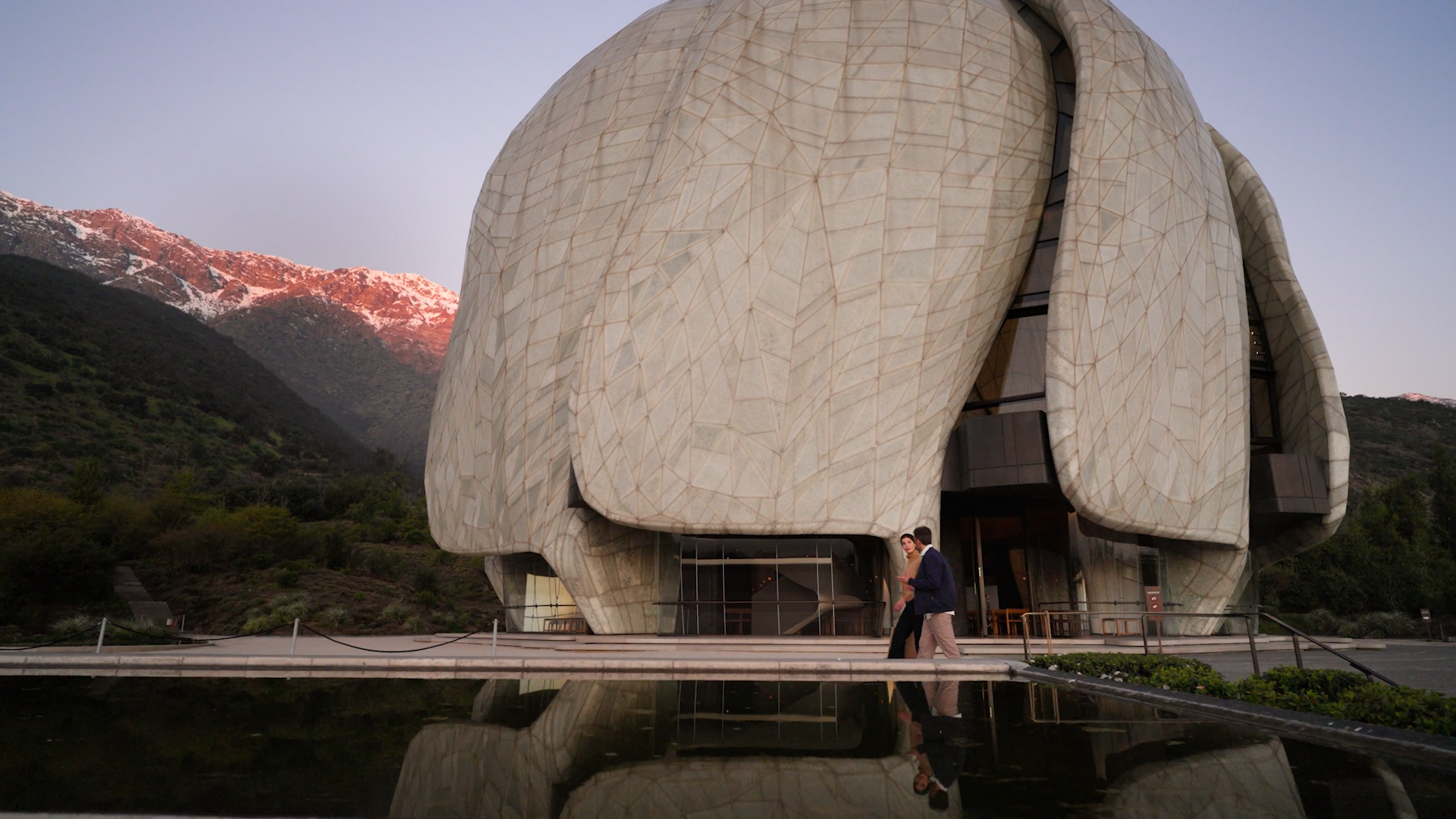 Modern building with a unique, organic shape and beige stone exterior, situated in a mountainous landscape with snow-capped peaks in the background, and two people walking near a reflecting pool in the foreground.