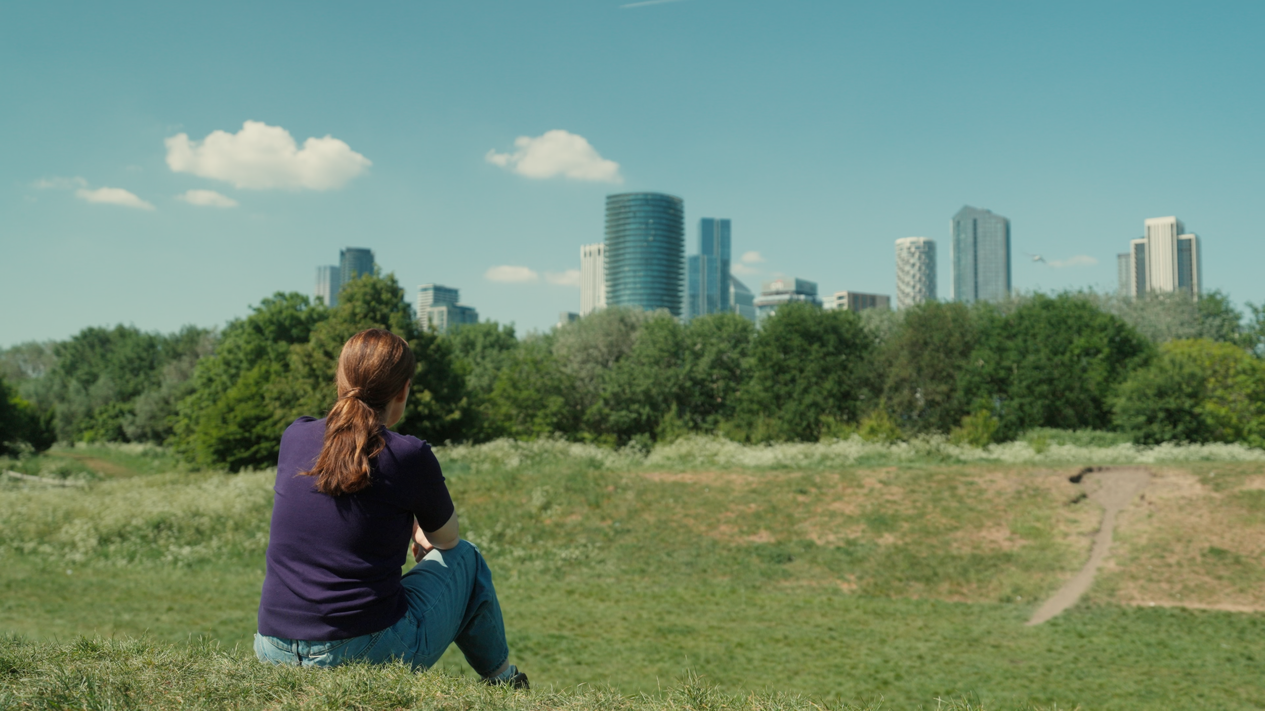 Person sitting on grass in a park, looking at city skyline with tall buildings and trees in the background on a clear, sunny day.