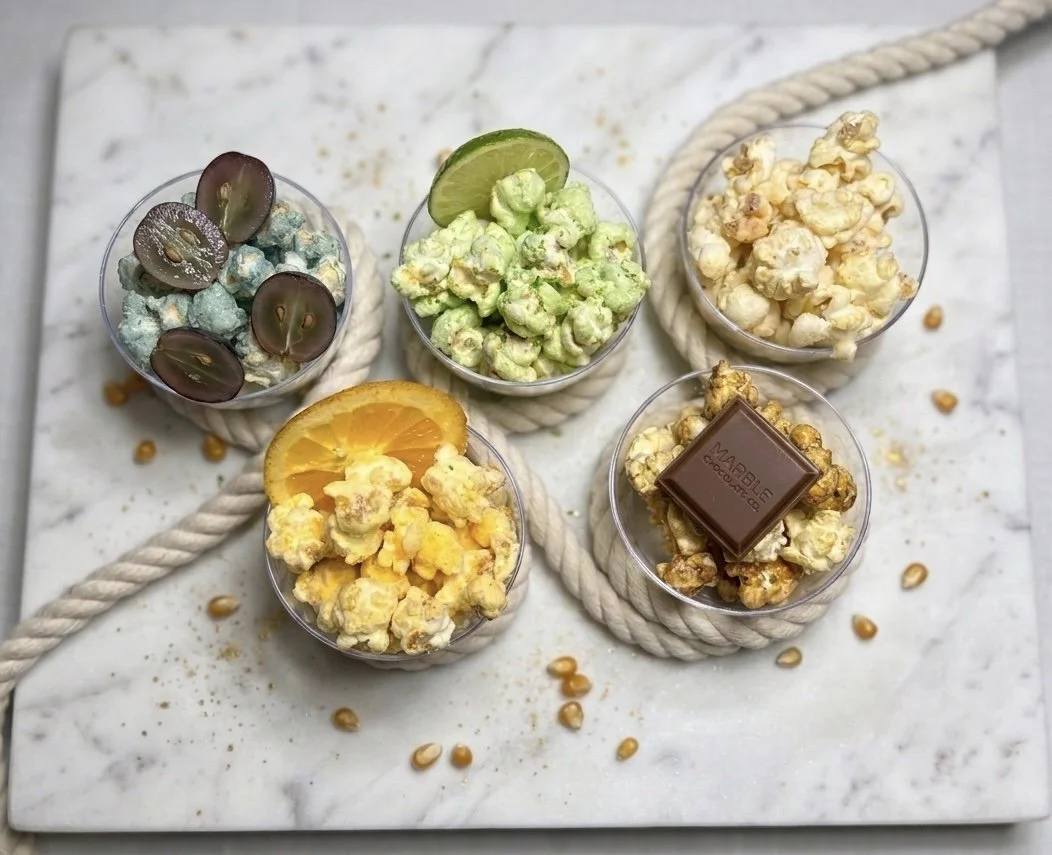 Five small glass bowls of popcorn with flavored toppings and garnishes, such as grapes, lime, orange, and a chocolate piece, on a marble surface with a thick rope.