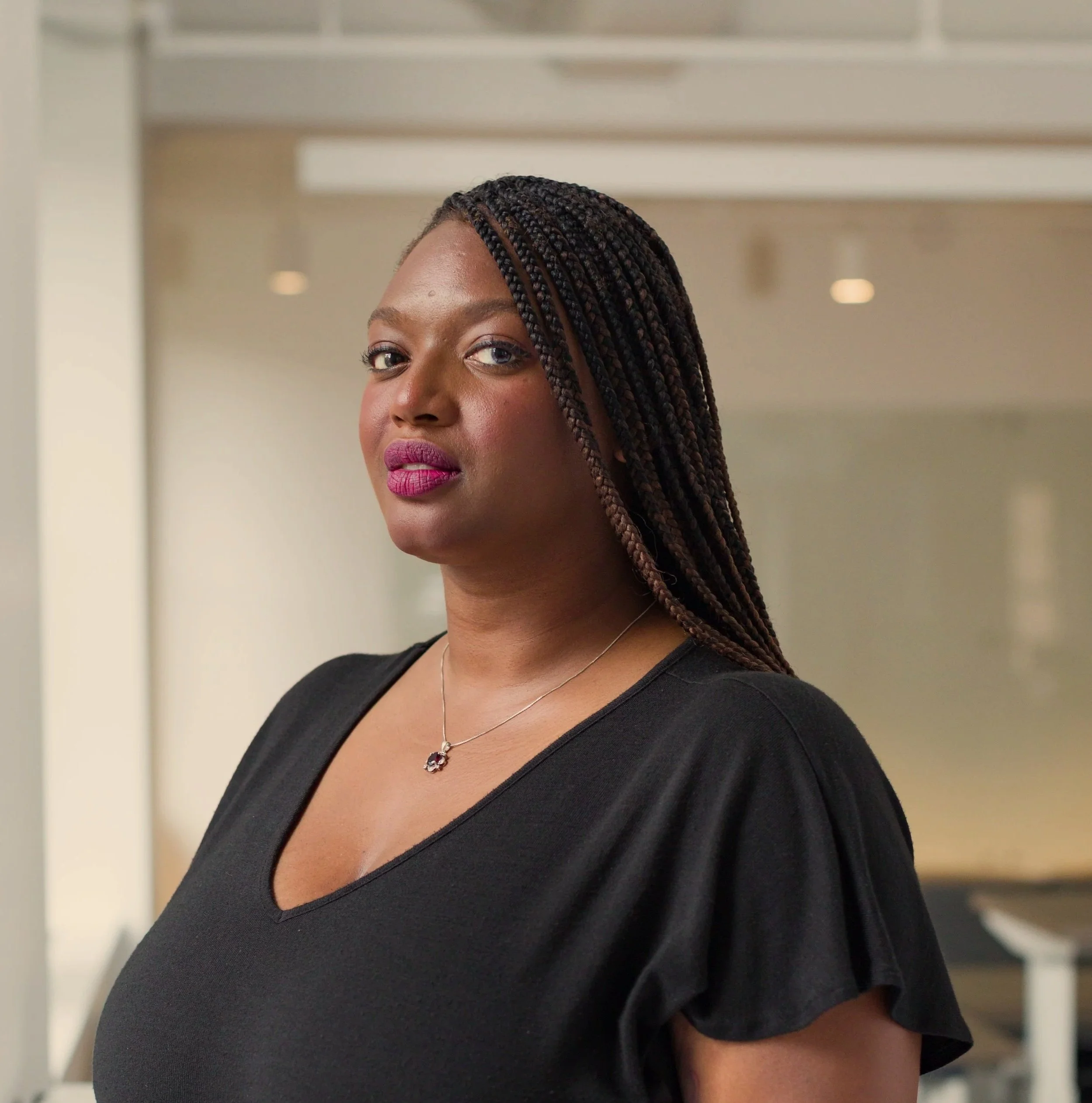A woman with long braided hair, wearing a black top and a necklace, looking confidently at the camera indoors.