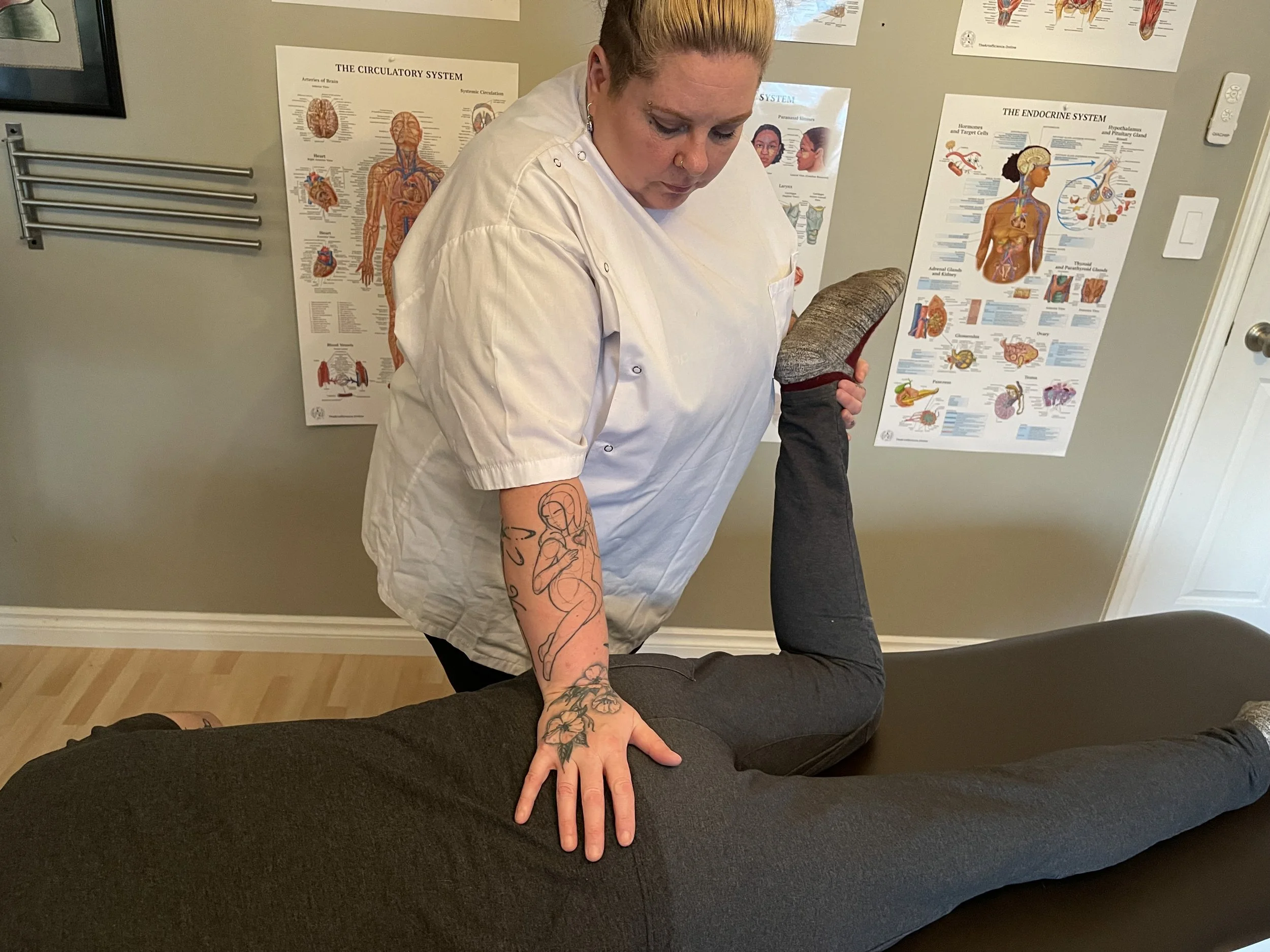 A person with tattoos on their arm performs a yoga pose called plank on a person lying face down on a yoga mat, in a room with medical posters on the wall.
