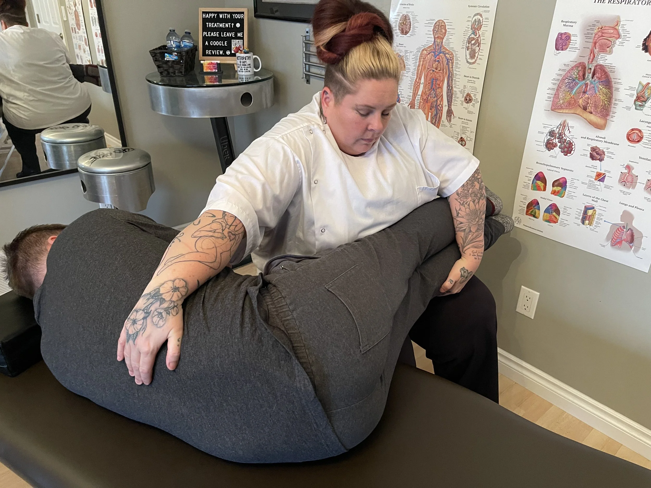A woman with tattoos in her arms and multicolored hair is lying face down on a massage table, receiving a massage in a room decorated with medical posters of human anatomy.